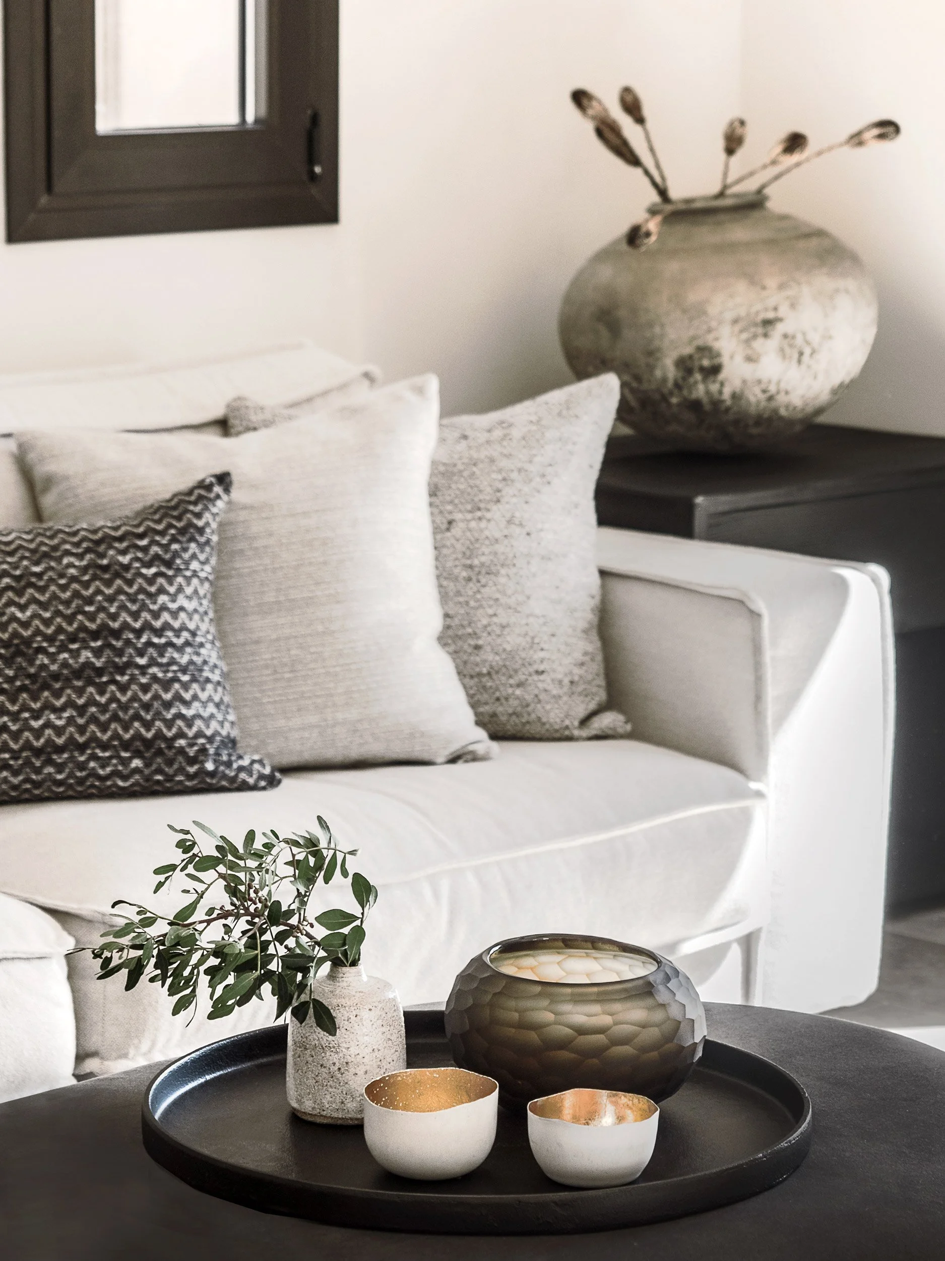 A round black tray with decorative ceramic vases, candles, and a small plant on a table in front of a sofa with beige and patterned pillows. A large ceramic vase with dried branches is in the background on a dark table near a white wall with a black 