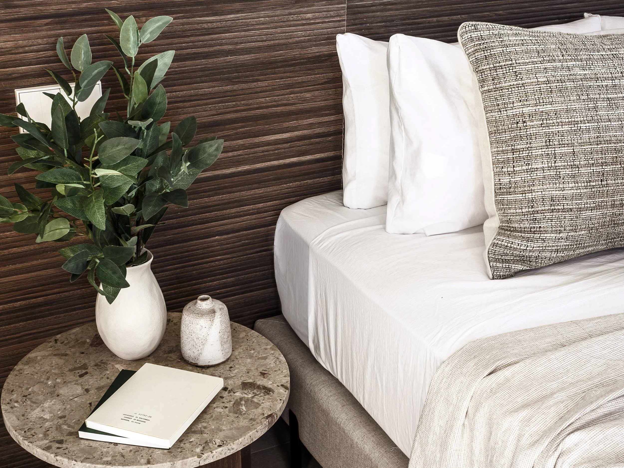 A bedroom corner with a wooden headboard, white bedding, decorative pillows, a plant in a white vase, and a marble-top side table with two notebooks and a small vase.