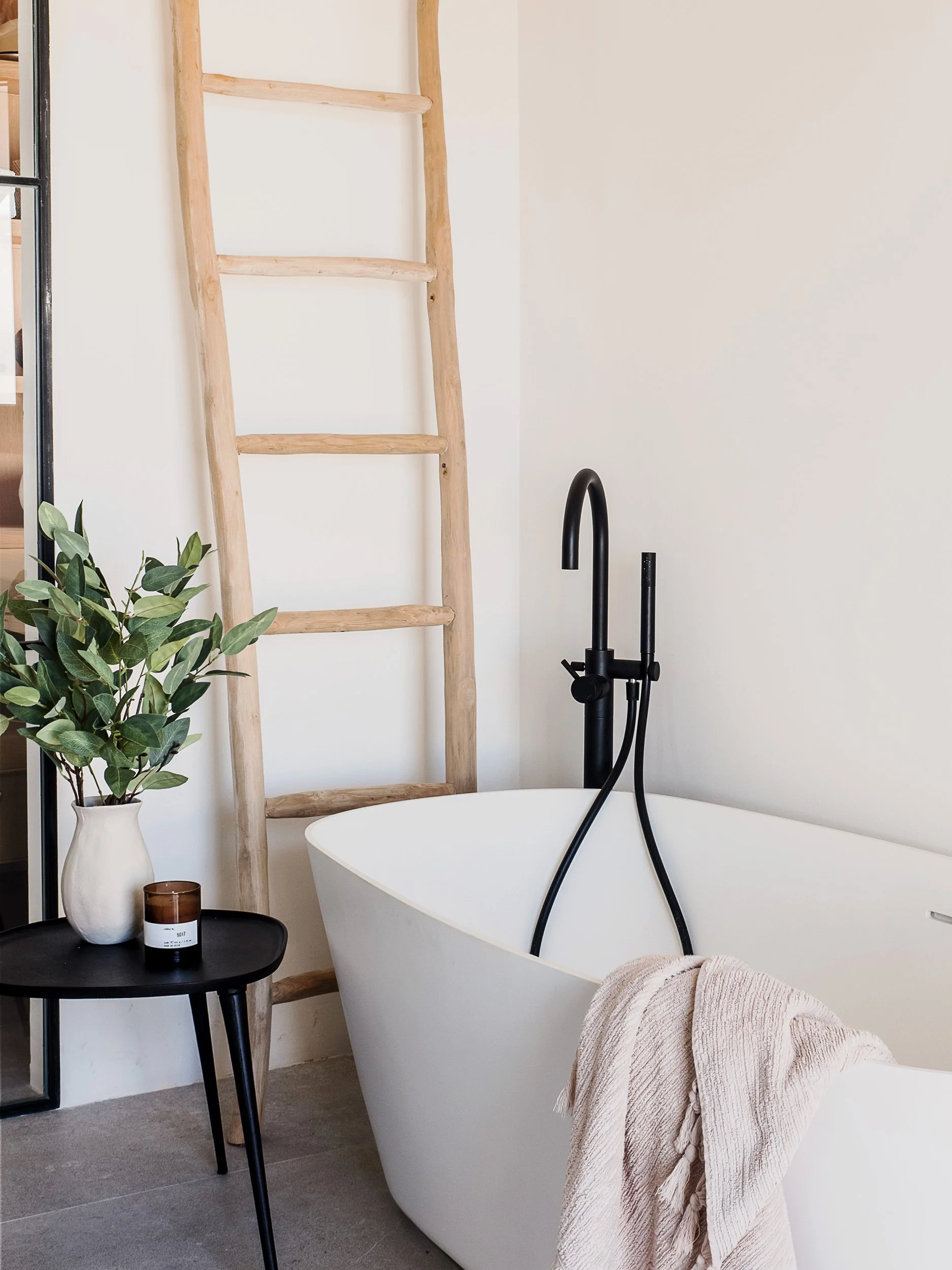 A bathroom scene with a white freestanding bathtub, black modern faucet, a beige textured towel draped over the edge, a small round black table holding a white vase with green leafy plant and a brown candle, to the left a wooden ladder leaning agains