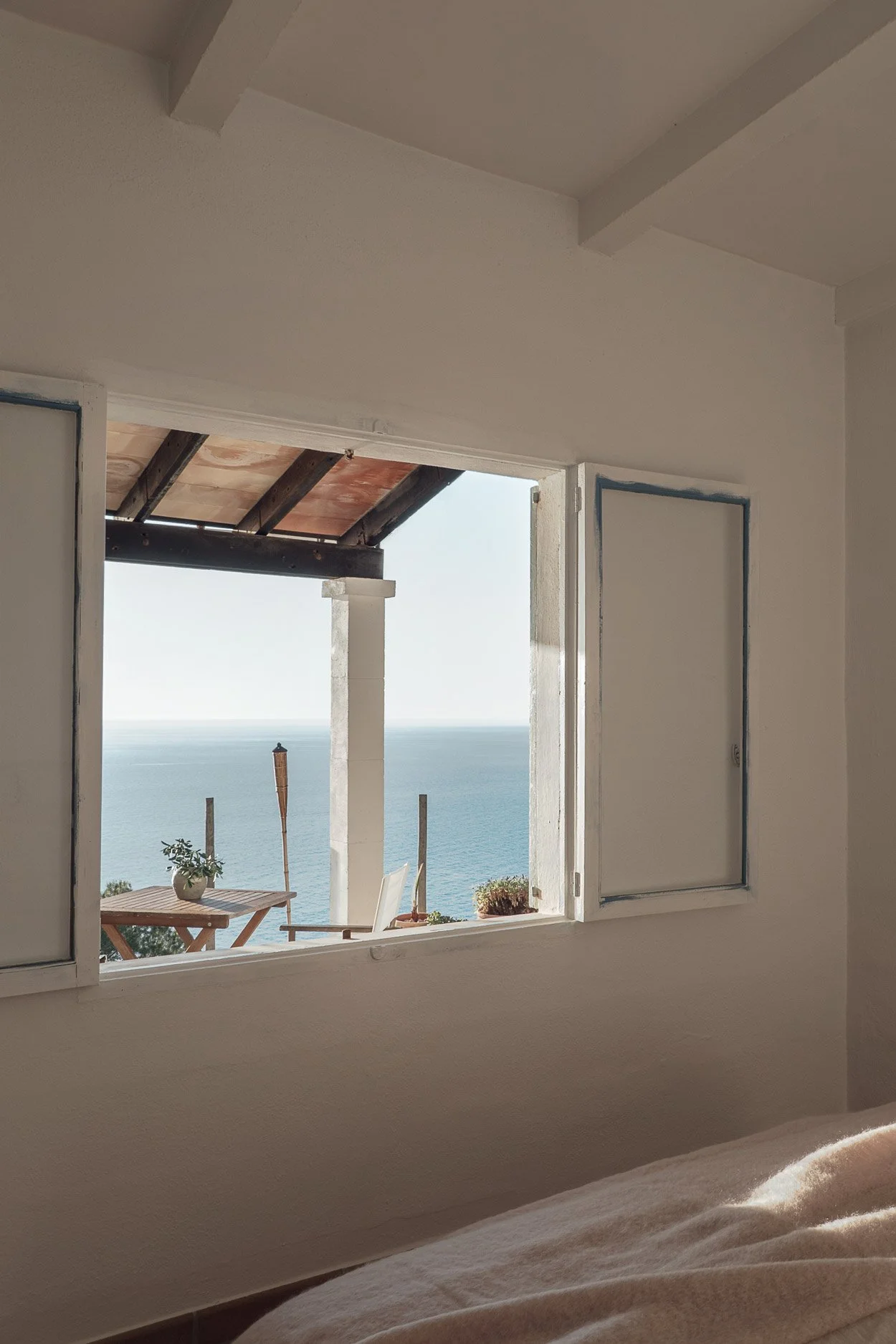 Interior of a room with an open window showing a balcony with a potted plant, outdoor table and chairs, and the ocean in the background.