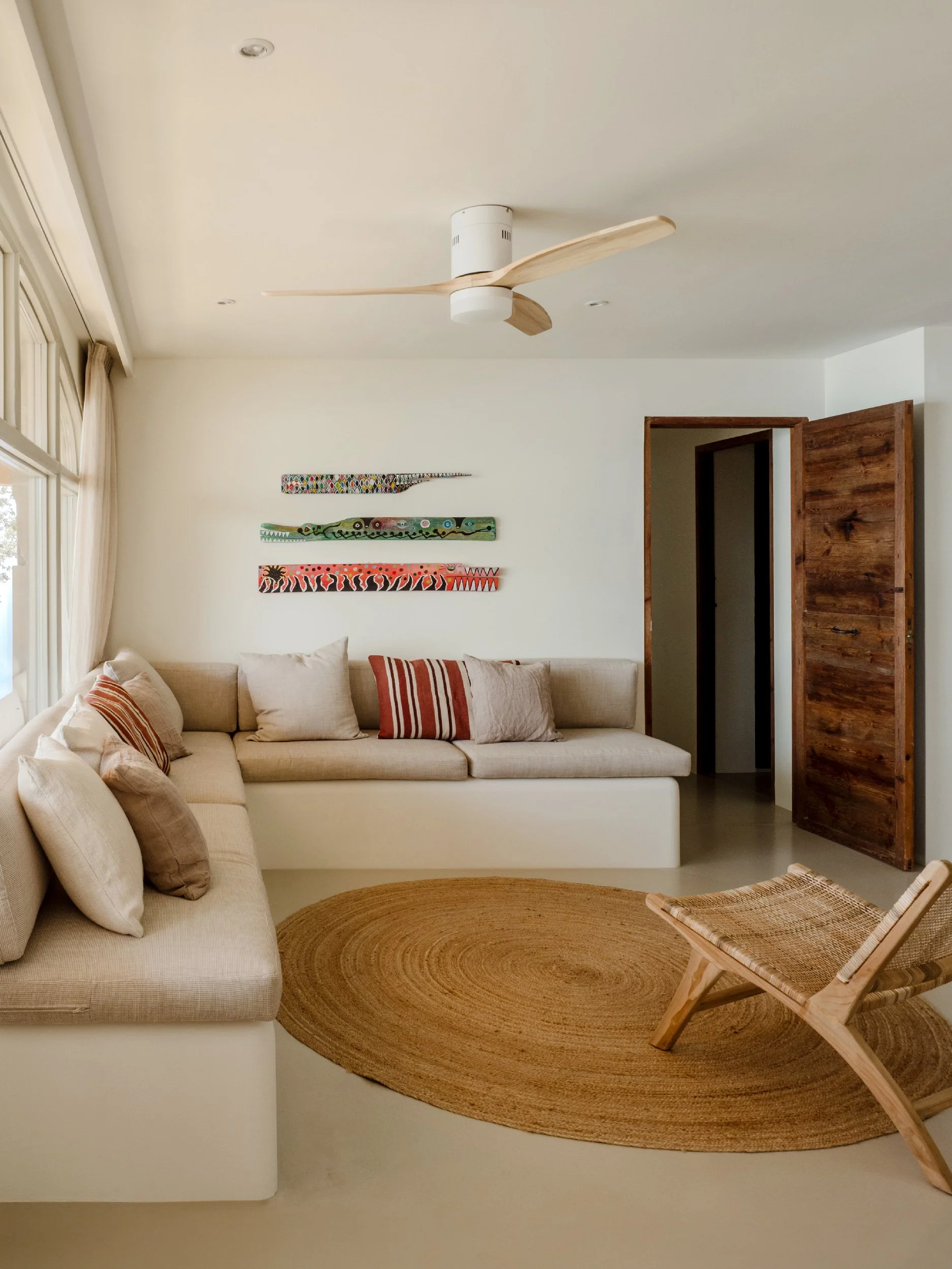 A living room with a beige sofa, throw pillows, a wooden lounge chair, a circular woven rug, a large window with curtains, a wall hanging with colorful patterns, a wooden sliding door, and a ceiling fan.