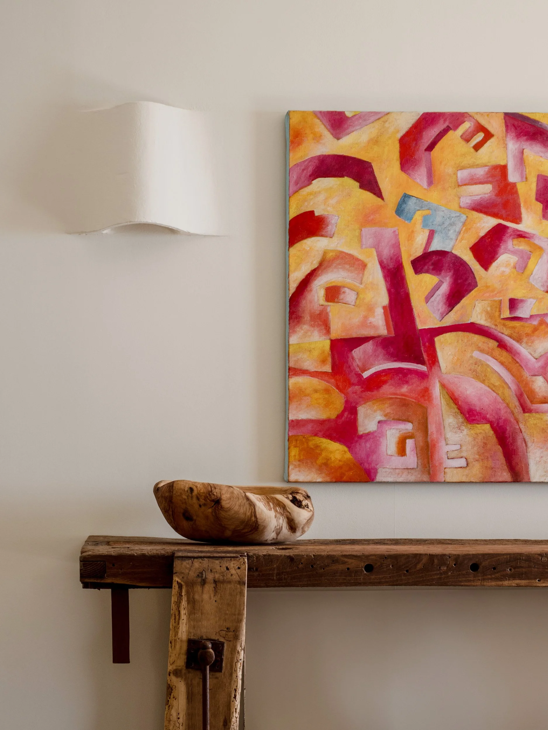 A white wall with a curved light fixture, a colorful abstract painting with red, yellow, and pink shapes, a carved wooden bowl, and a rustic wooden table.