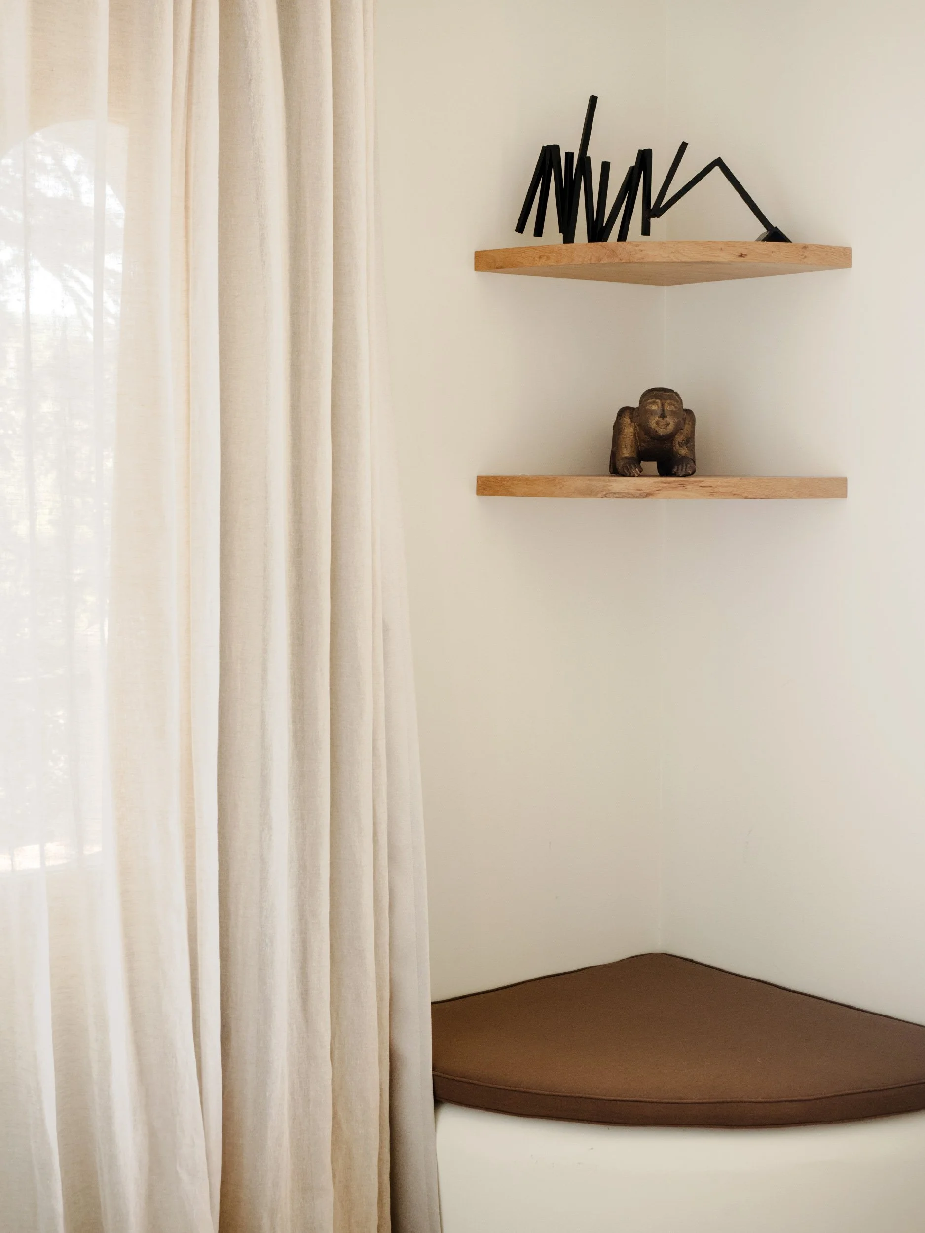 A corner of a room with beige curtains on the left, two wooden shelves on the wall, one with black sculptural decorations and the other with a small figurine resembling a bear. The floor is brown.