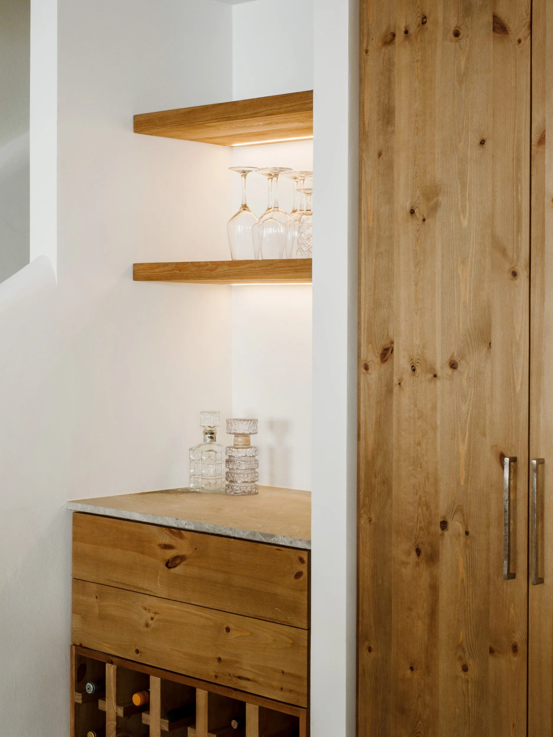 A wooden cabinet with open shelving above a countertop, displaying glassware, and a wine rack below with wine bottles.