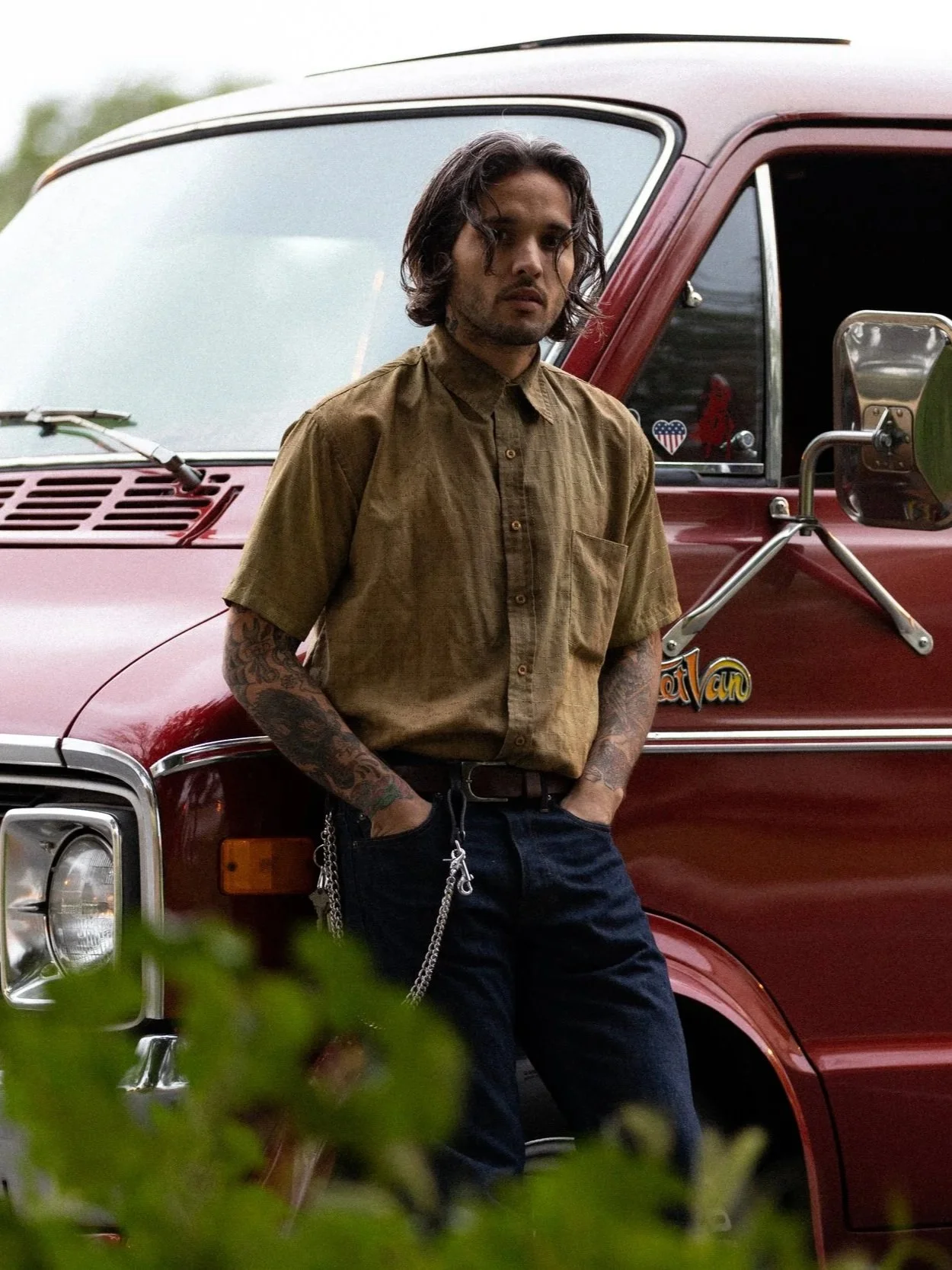 A young man with tattoos and wavy dark hair standing in front of a red van, wearing a brown button-up shirt and dark pants, with his hands in his pockets.