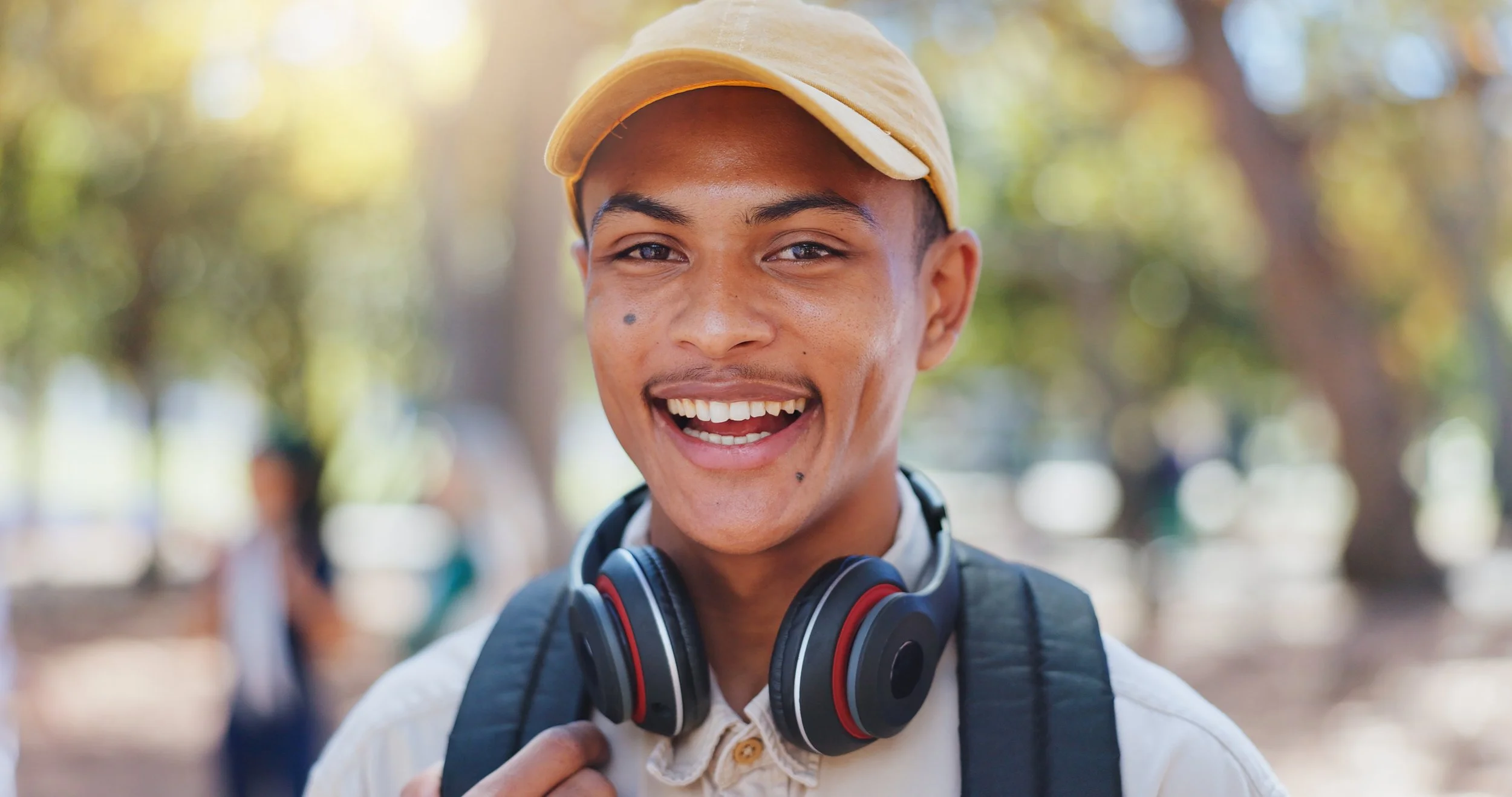 A young man smiling outdoors, wearing a beige cap, headphones around his neck, and a backpack