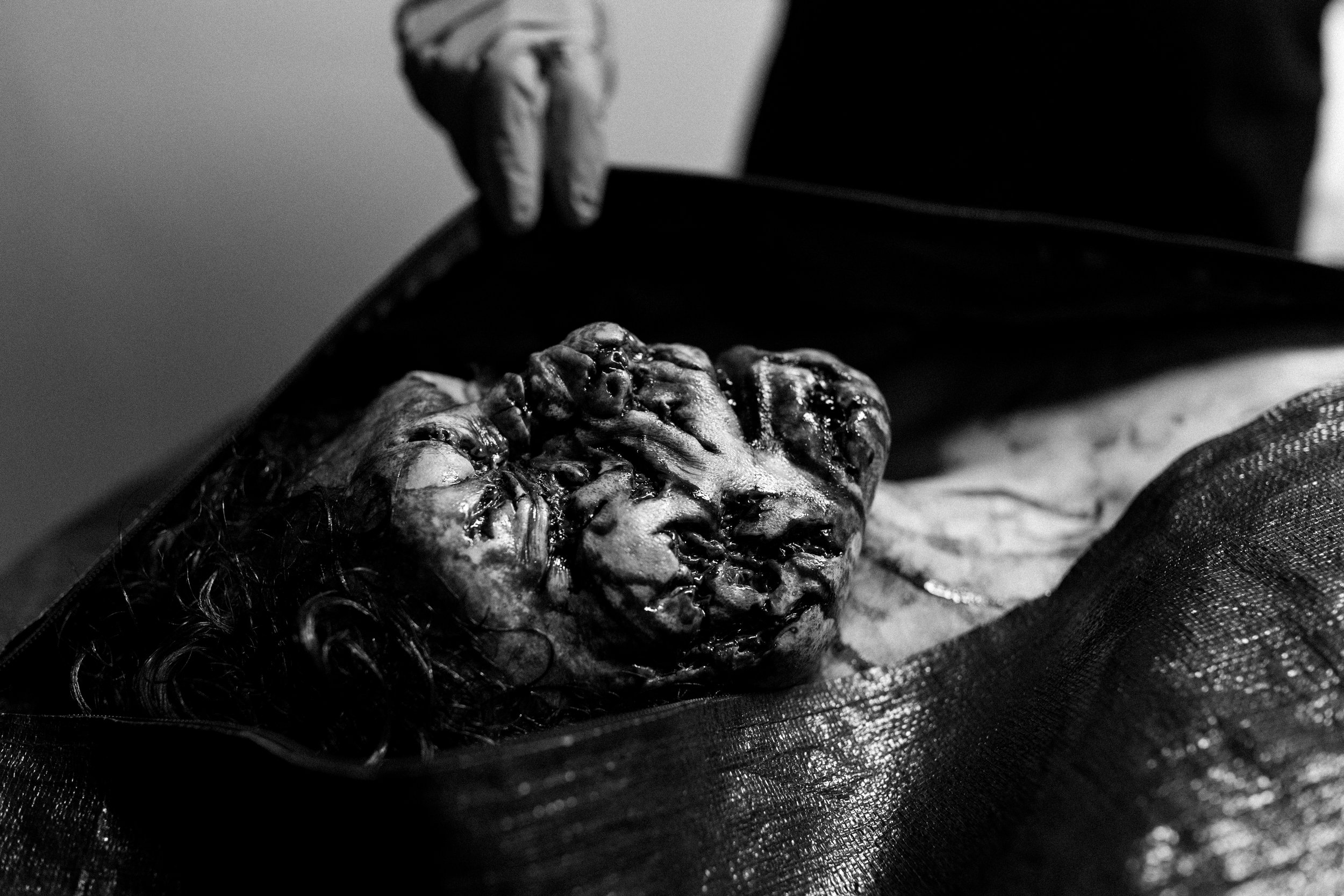 A person wearing gloves is holding a black bag containing a preserved human head with visible features, hair, and details in black and white.