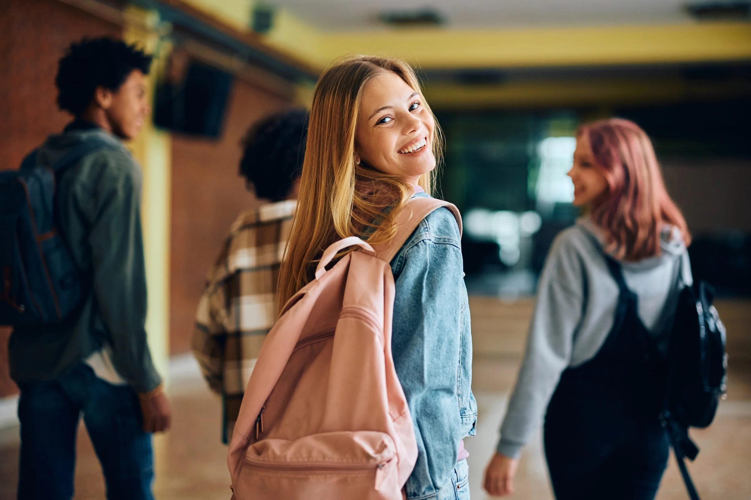 A smiling girl with long blonde hair, wearing a denim jacket and pink backpack, looking at the camera in a school hallway with other students in the background.