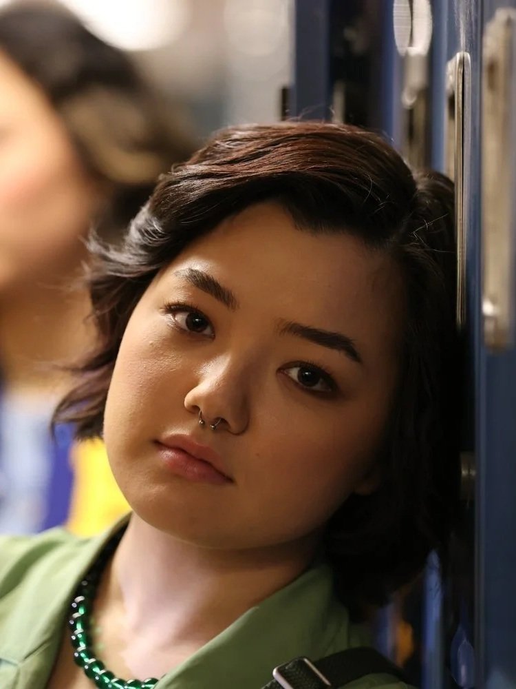 Person with short dark hair, wearing a green shirt, green beaded necklace, and a septum piercing, leaning against a locker in a school hallway.