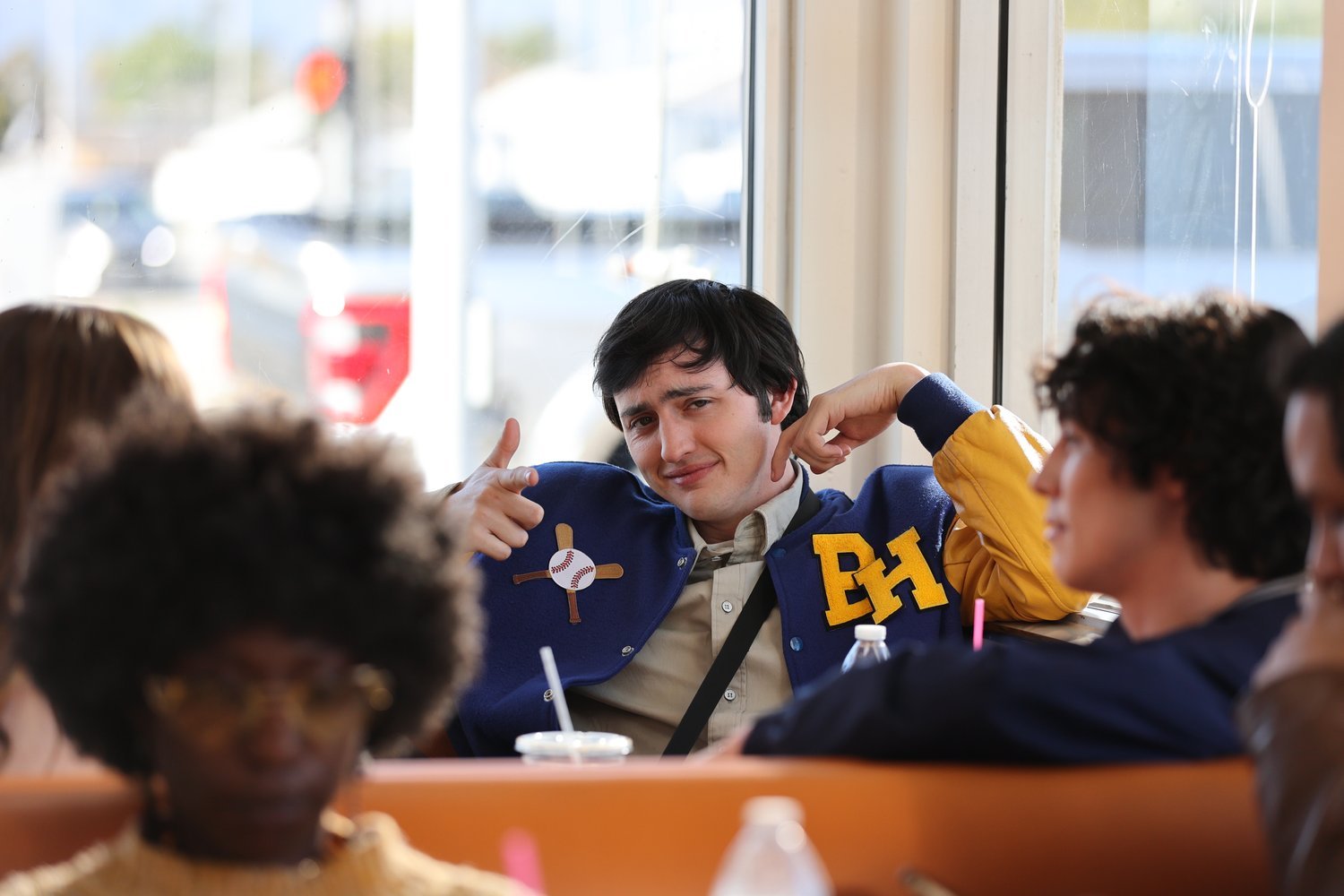 A young man with dark hair making a playful gesture with his fingers, wearing a blue letterman jacket with a yellow 'EH' patch, sitting indoors with others around, near a window with a parking lot outside visible.
