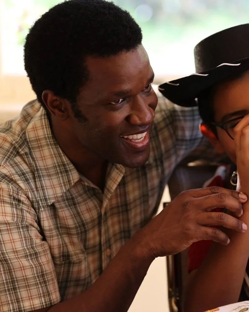 A man with black curly hair smiling and engaging with a young person wearing glasses and a black hat, in an indoor setting with natural light.