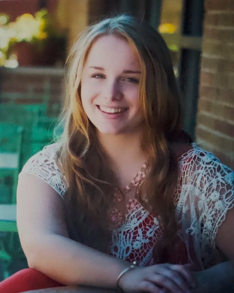 A young woman with long, wavy brown and blonde hair smiling outdoors, wearing a white lace top and a pink necklace.