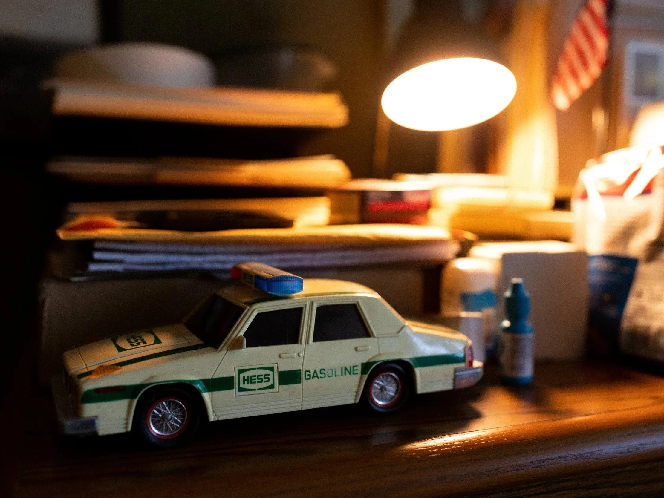 Toy police car with Hess Gasoline logo on a wooden surface, with bottles, papers, and a lamp in the background.