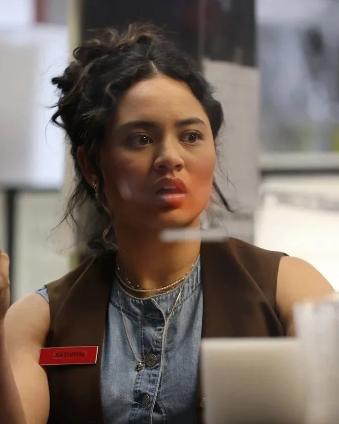 A young woman with dark curly hair and earrings working at a computer in a workplace, wearing a brown vest over a denim shirt with a red name tag that says 'Kathryn' and a silver necklace.