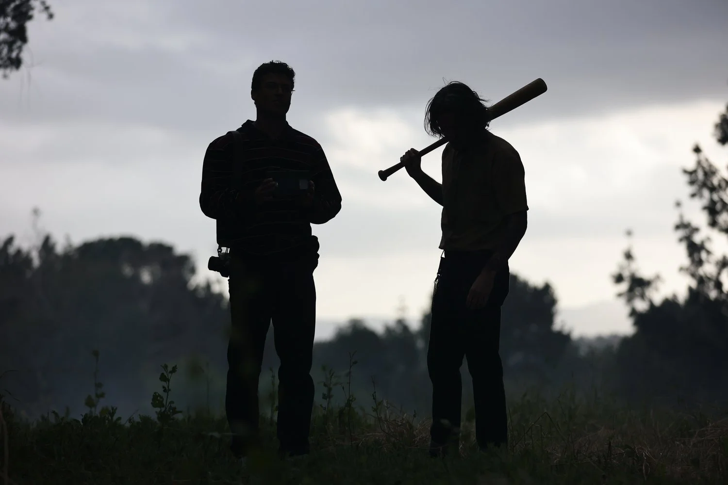Silhouettes of two men, one holding a baseball bat and the other holding a camera, standing outdoors against a cloudy sky with trees in the background.