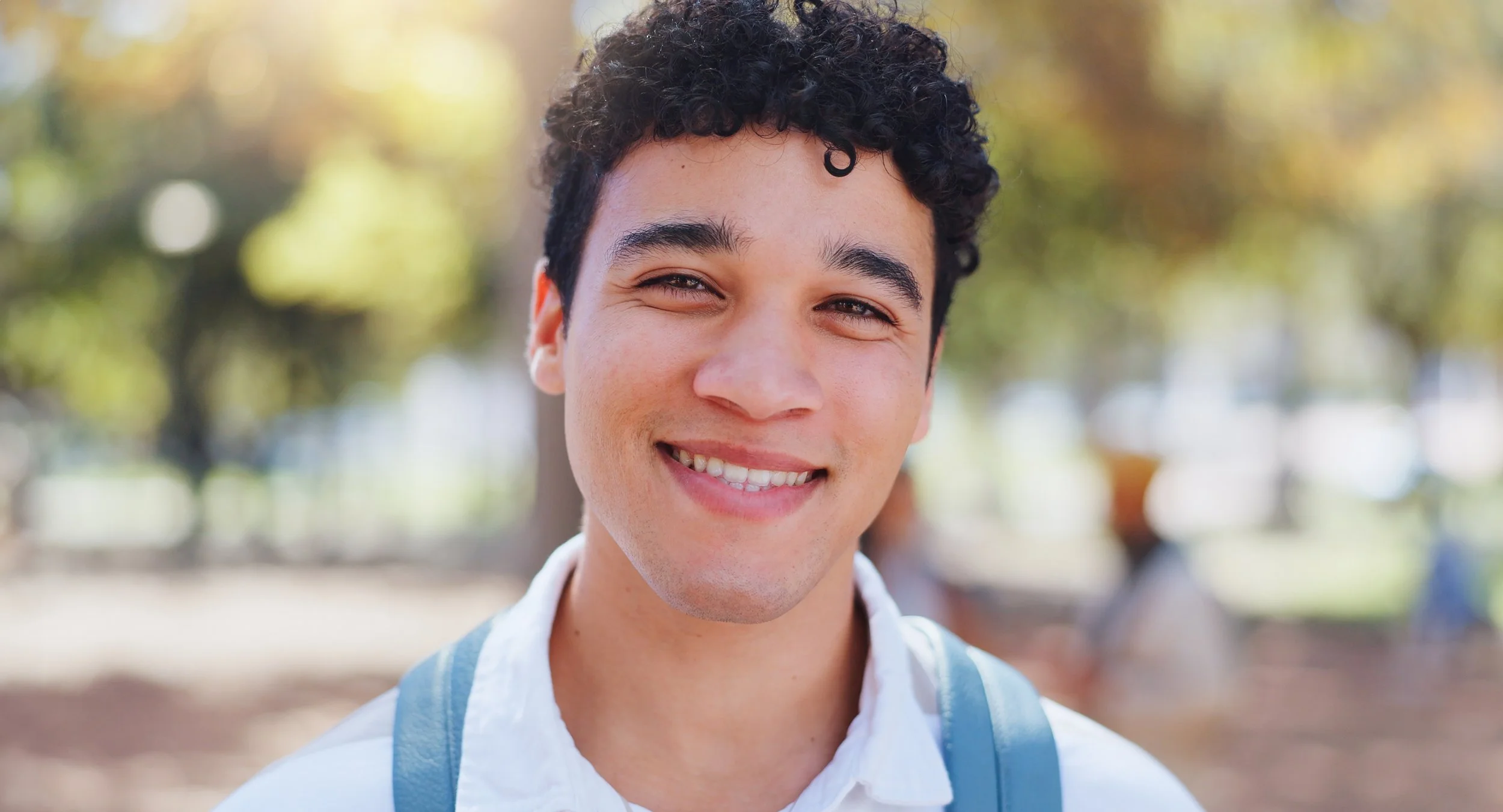 Smiling young man with curly dark hair, wearing a white shirt and a backpack, outdoors with blurred trees and people in the background.