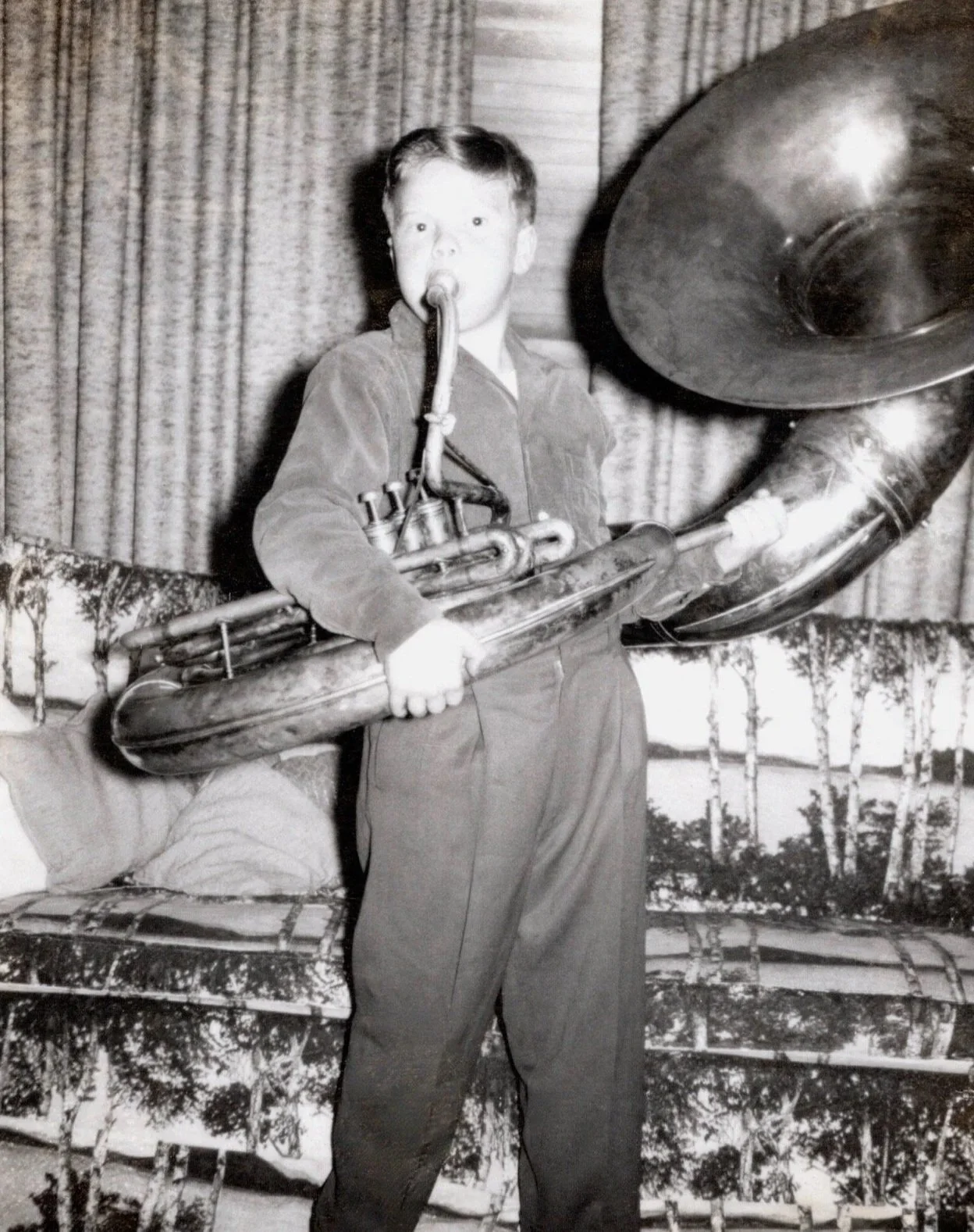 A young boy playing a large bass saxophone in a room with a curtain background and a nature scene mural or backdrop. The boy has short hair and is wearing a long-sleeved shirt and pants.