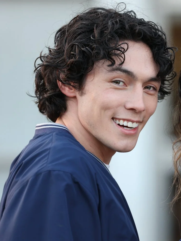 Young man with curly dark hair smiling and wearing a blue graduation gown.