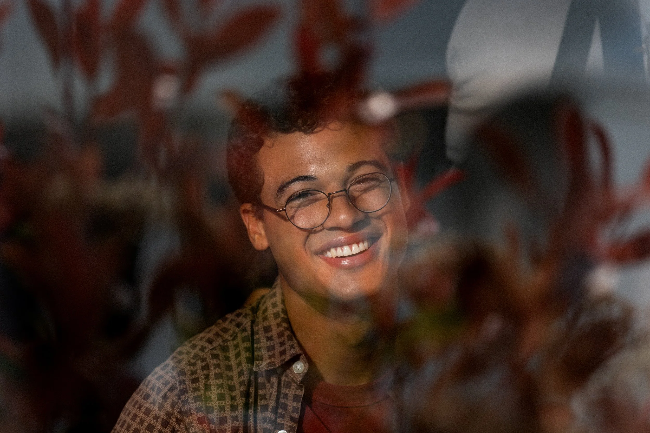 A person with glasses and curly hair smiling behind reddish-brown leaves.
