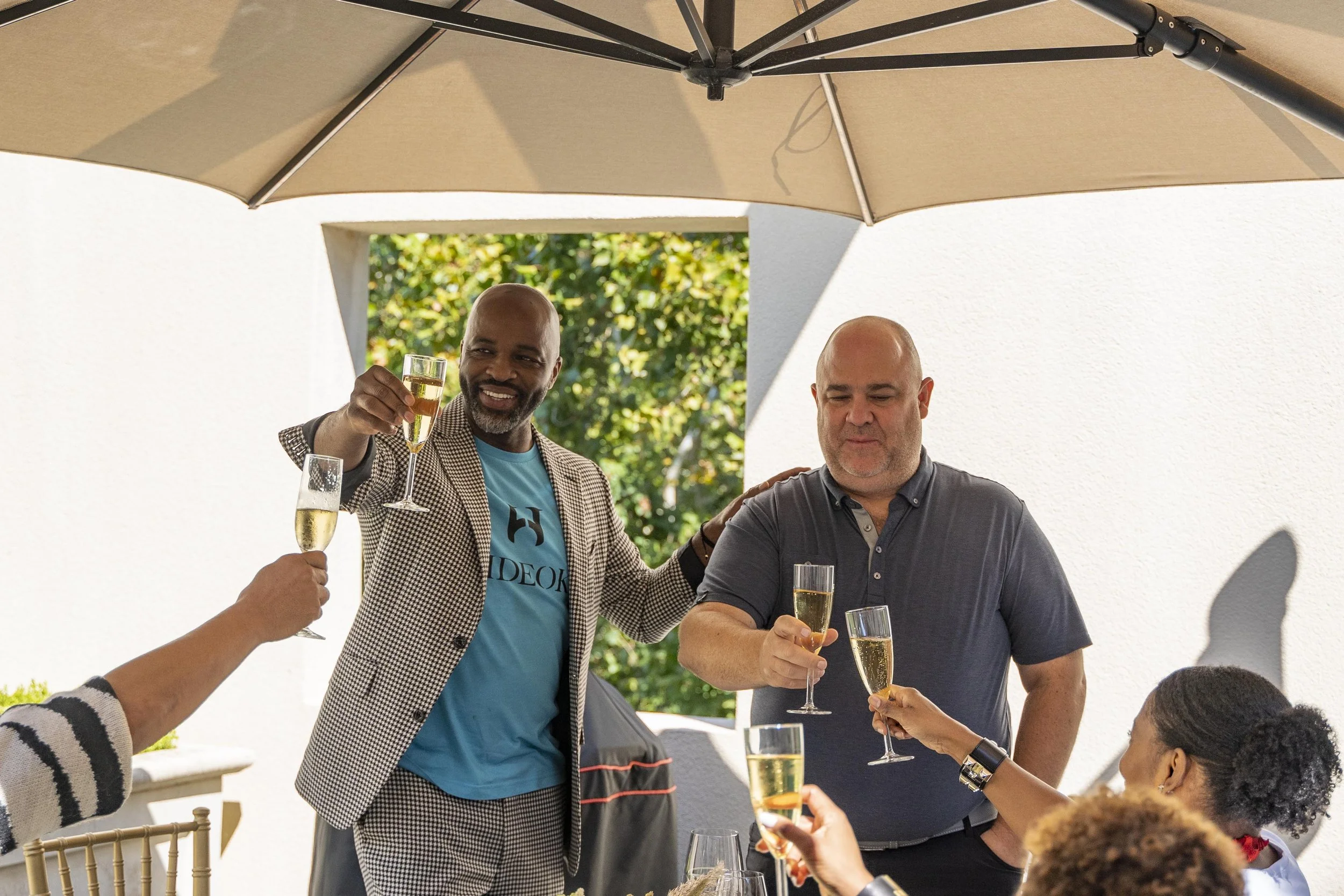 People at an outdoor gathering raising glasses of champagne in a toast under a beige patio umbrella.