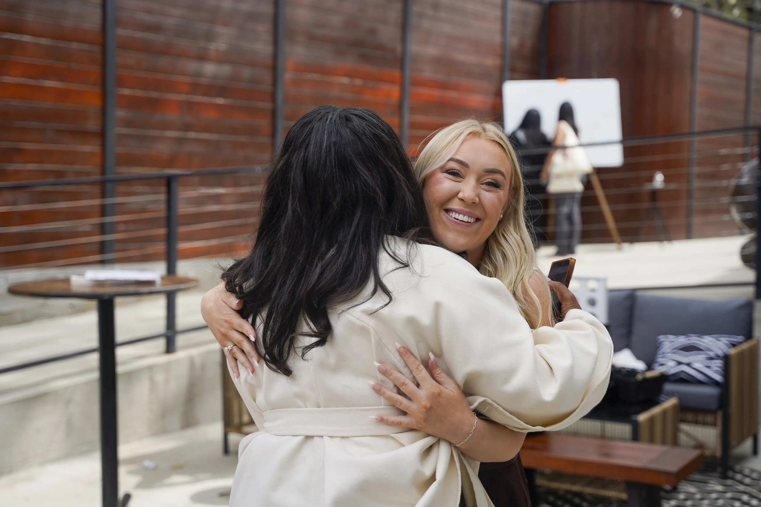 Two women hugging each other and smiling, in an outdoor area with wooden fencing, a table, and chairs.