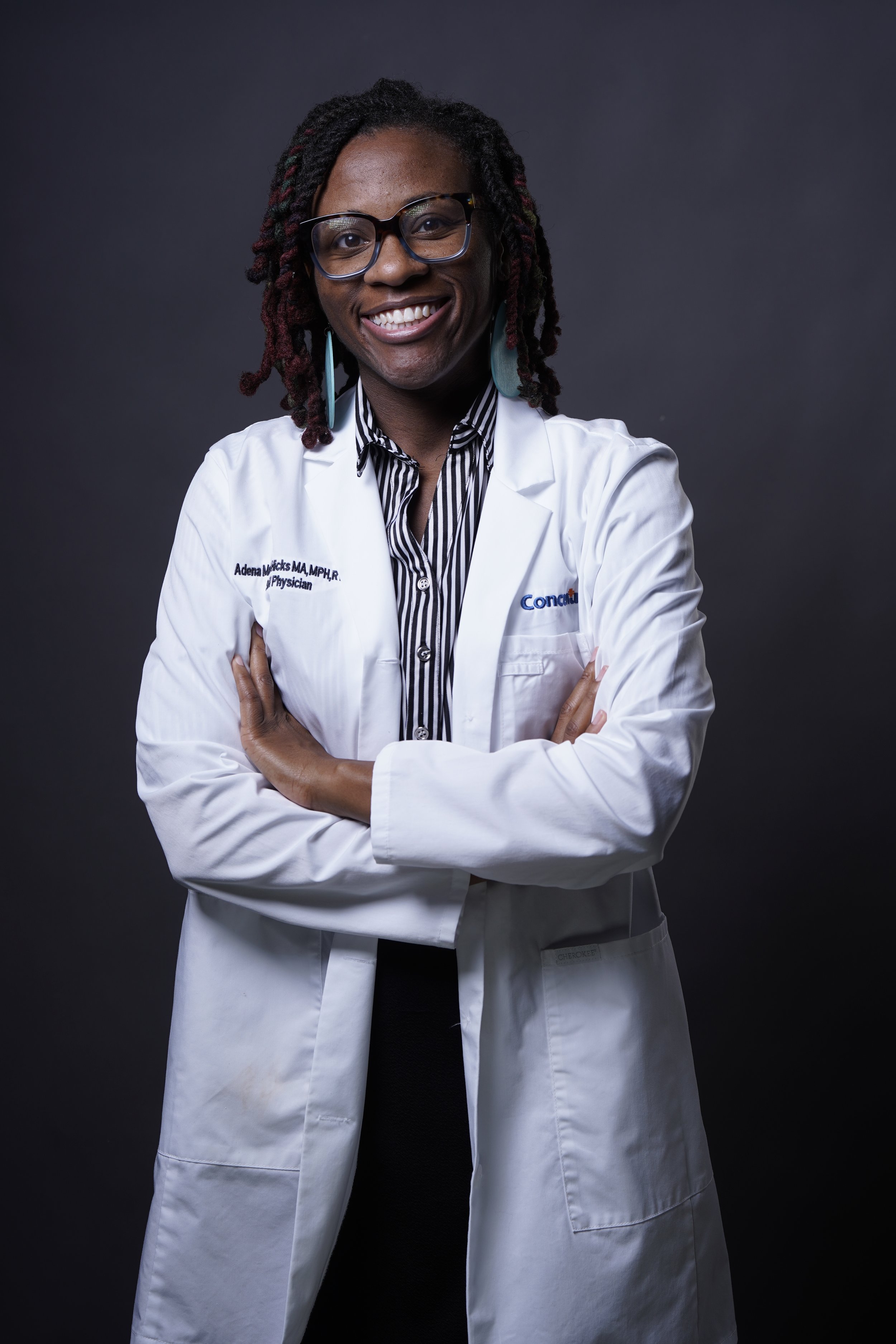 A confident African American woman with dreadlocks, wearing glasses, a white medical coat, and a striped shirt, standing with arms crossed against a dark gray background.