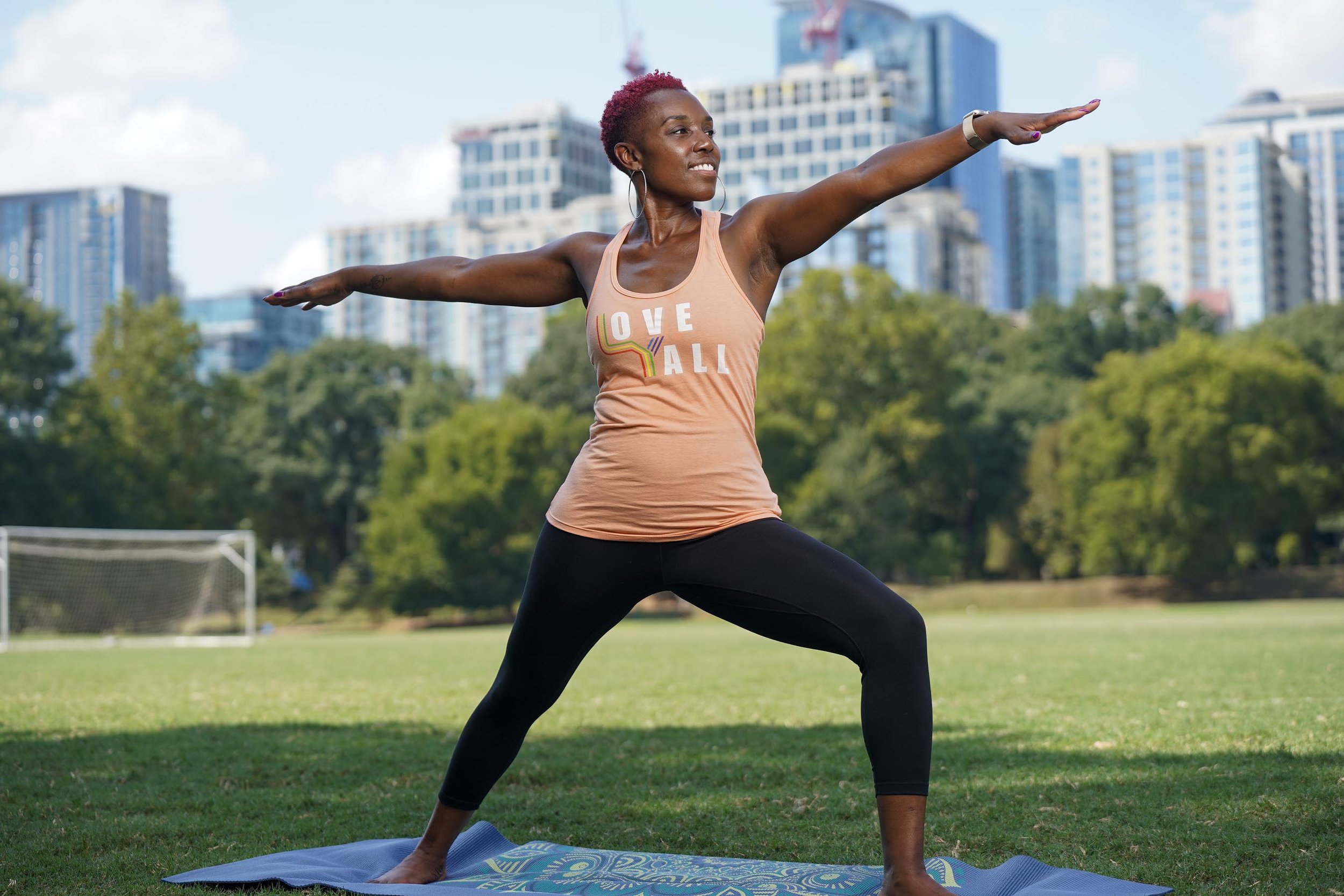A woman with short red hair practicing yoga outdoors on a blue mat in a park, with trees and tall buildings in the background, wearing a peach tank top and black leggings.