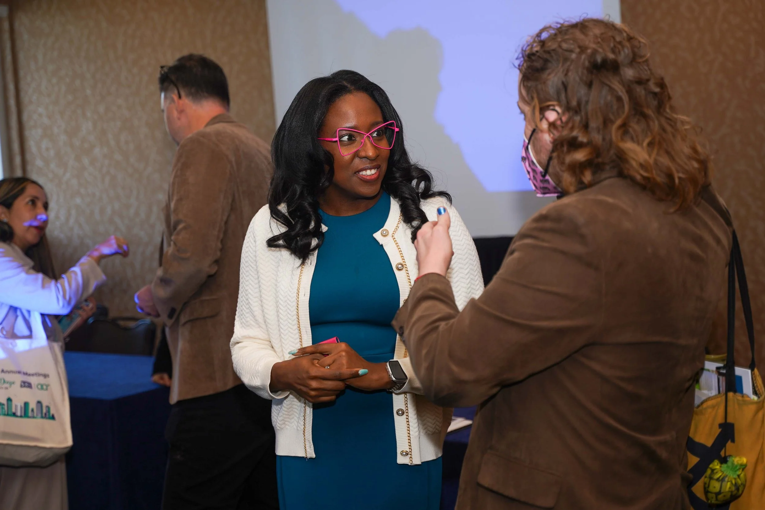 Two women engaged in conversation at a conference, with a third person in the background and a presentation screen.