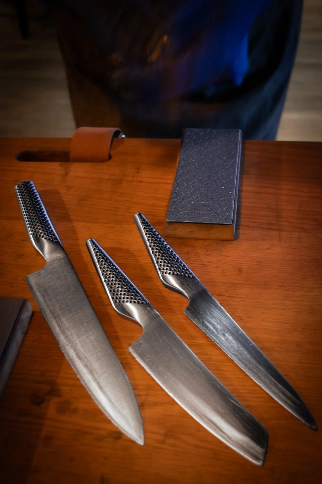 Three Japanese-style knives with perforated handles and a sharpening stone on a wooden table.
