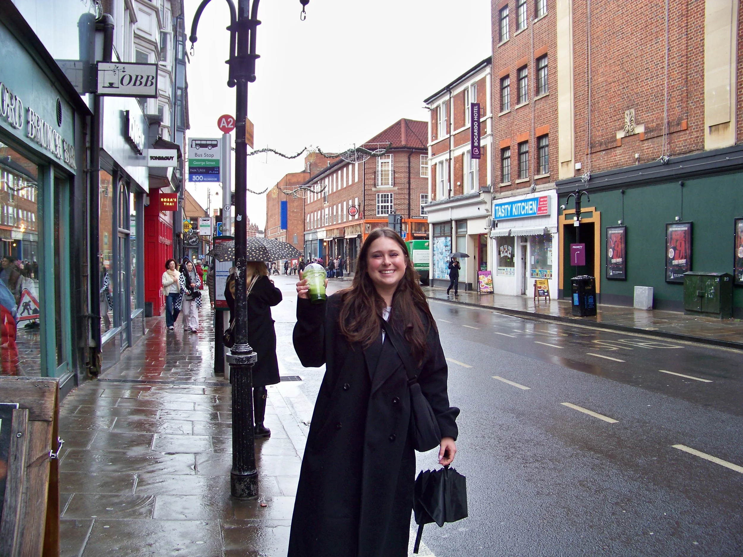 A smiling young woman with brown hair standing on a city sidewalk holding a green beverage. She is wearing a black coat, and there are shops, pedestrians, and brick buildings on a rainy day.