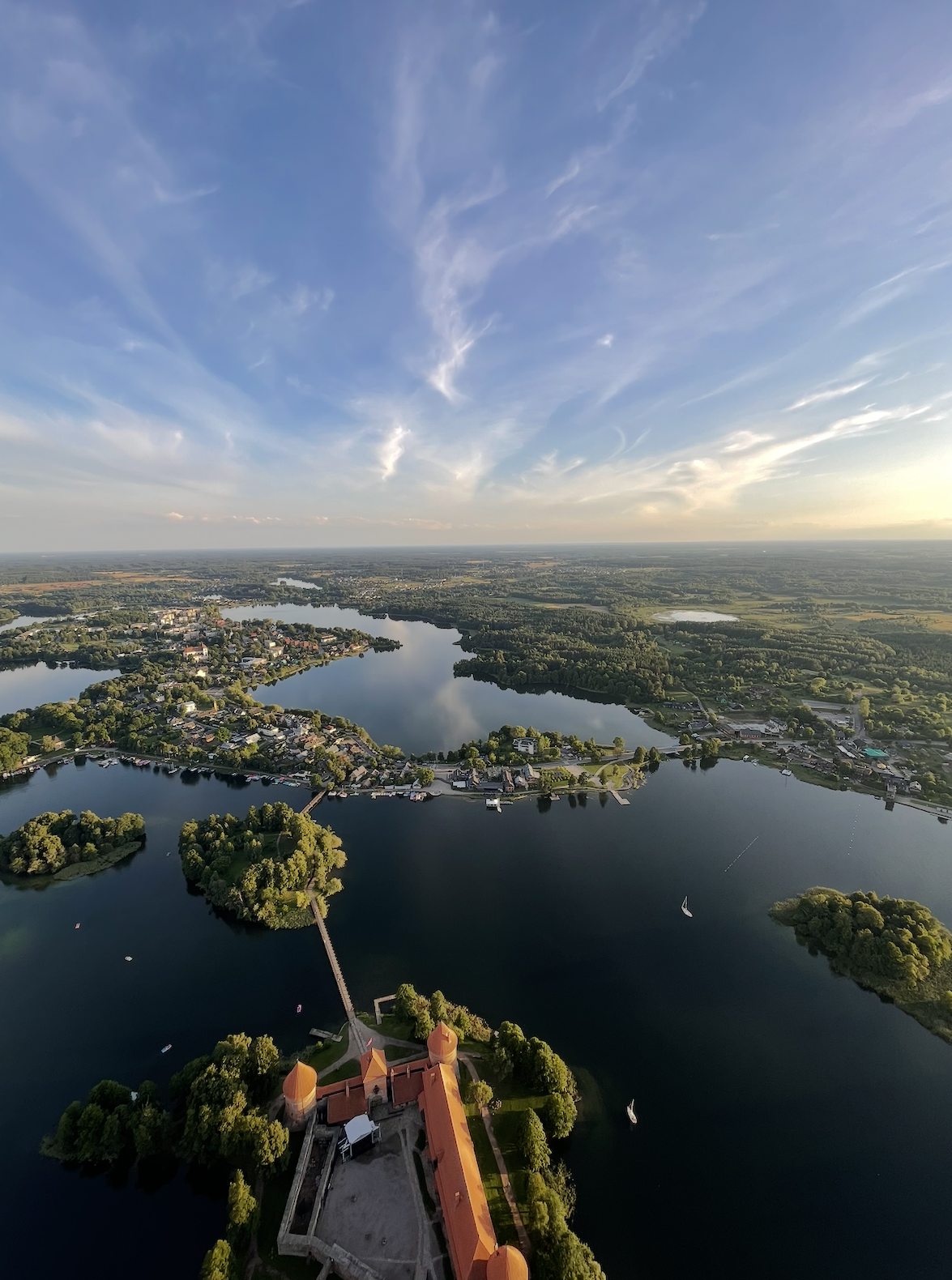 Aerial view of a large lake with surrounding green trees, houses, and a small castle or historic building with orange rooftops in the foreground, under a sky with wispy clouds.