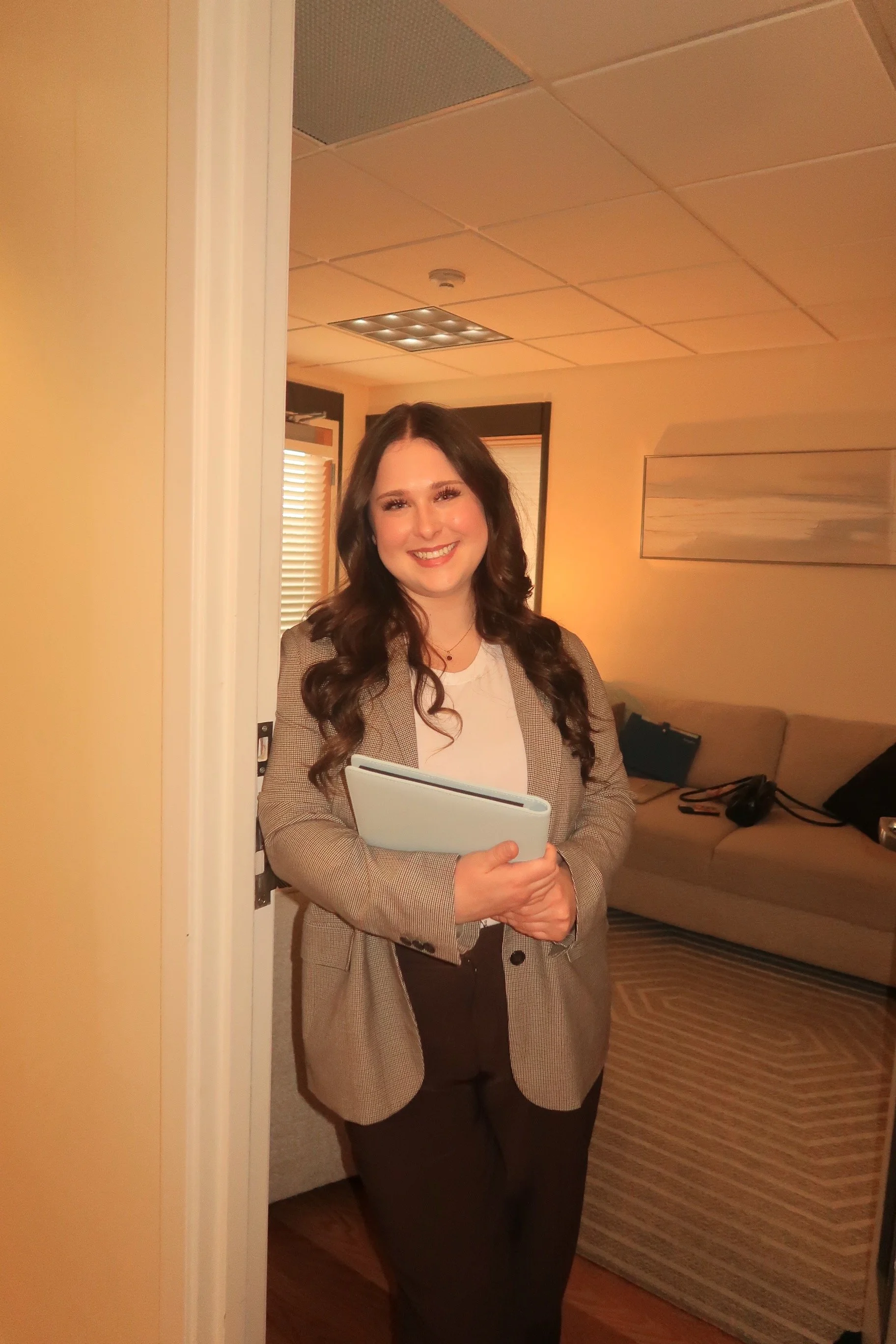 A woman with long, dark, curly hair is standing in a room, smiling at the camera. She is dressed in a beige blazer and dark pants, holding a white tablet or folder in her left hand. The room has a beige couch, a window with blinds, and a piece of artwork on the wall.
