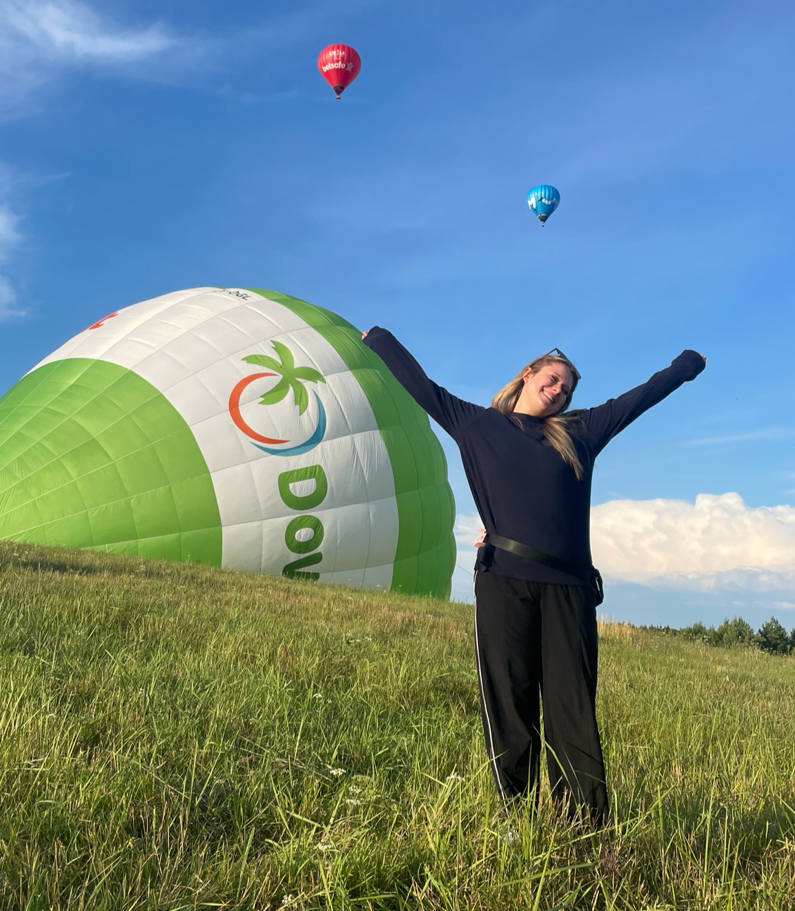 A woman celebrating outdoors with a large green and white hot air balloon on the ground and two smaller hot air balloons in the sky.