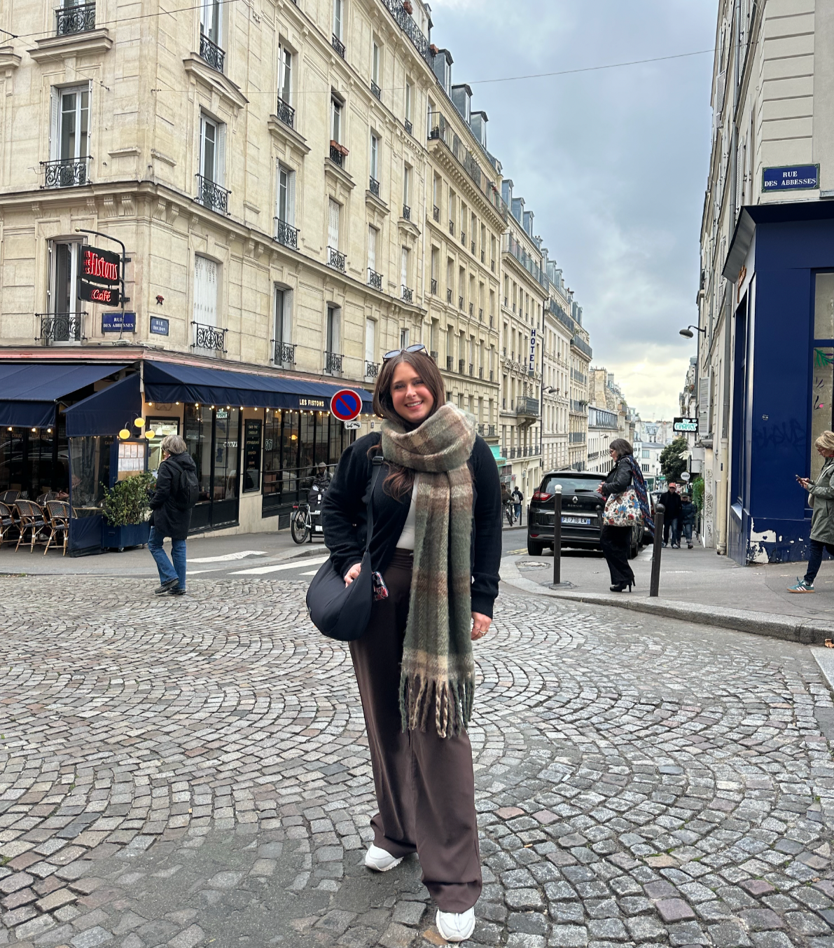 A woman with long brown hair, wearing a black jacket, beige scarf, and brown pants, stands on a cobblestone street in Paris, France, smiling at the camera. The street features classic Parisian architecture with cream-colored buildings, a restaurant with outdoor seating, and several pedestrians walking by.