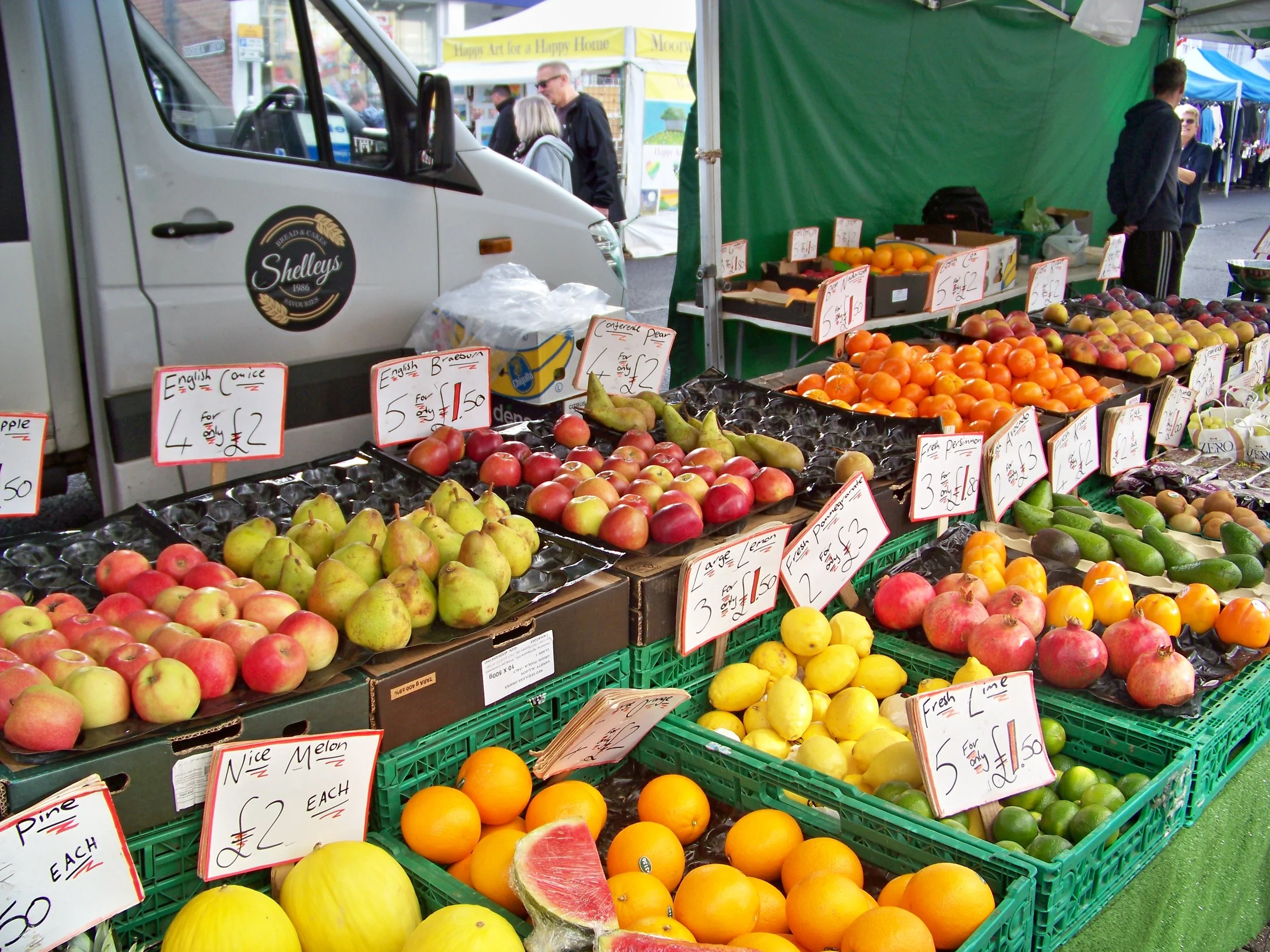 A street market stall with various fresh fruits including apples, oranges, pears, and watermelons, with price signs displayed. Behind the stall, people are shopping and walking near tents.