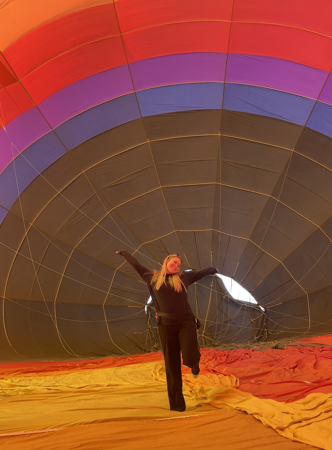 A woman standing inside a deflated hot air balloon with arms outstretched and smiling, surrounded by colorful fabric in hues of orange, red, purple, and black.