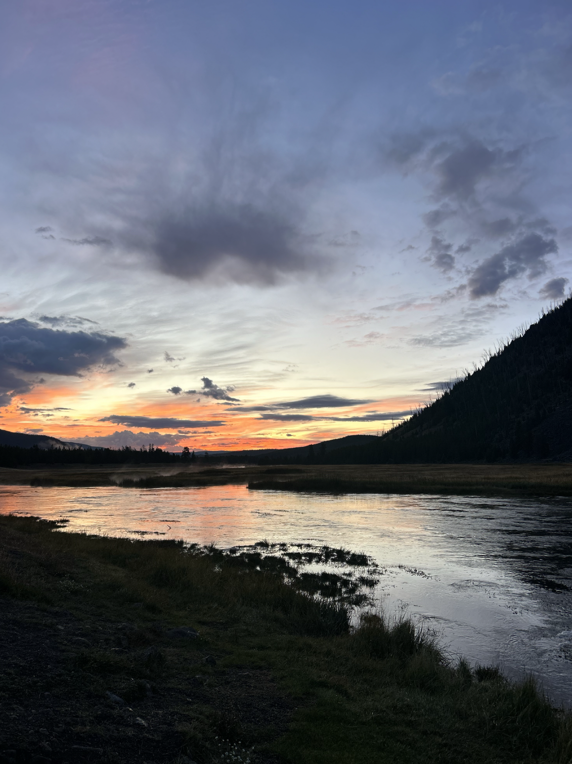 A scenic view of a sunset over a river with mountains and a partly cloudy sky.