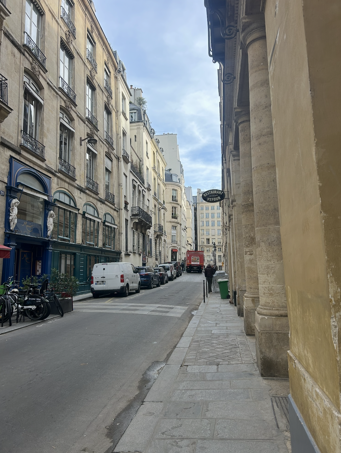 A narrow city street with parked cars and bicycles, surrounded by classic European-style buildings with balconies and ornate details. There is a sidewalk on the right side with a green trash bin, and a person walking away in the distance. A sign for a restaurant named 'Vefour' hangs from a building on the right.