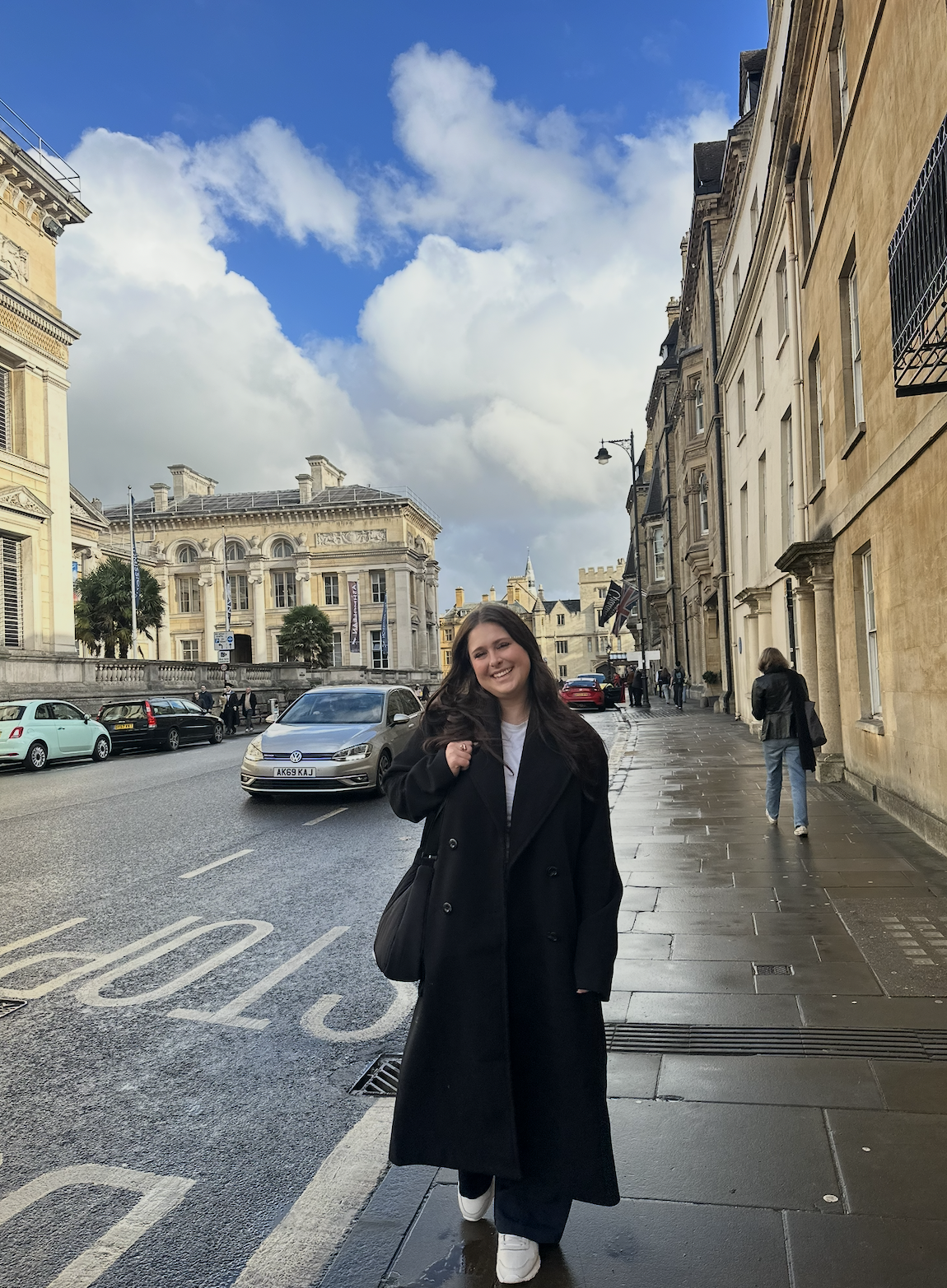 A young woman with long dark hair smiling and walking on a wet city sidewalk, wearing a long black coat, white sneakers, and carrying a black bag. The background features historic buildings, parked cars, and a partly cloudy sky.