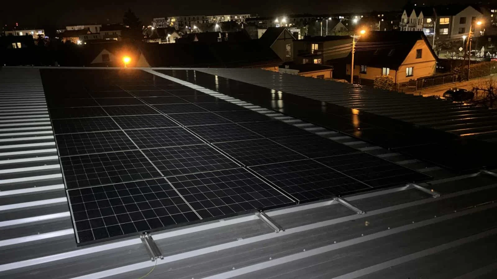 Nighttime view of solar panels installed on a building's roof, with a cityscape and houses illuminated by streetlights in the background.