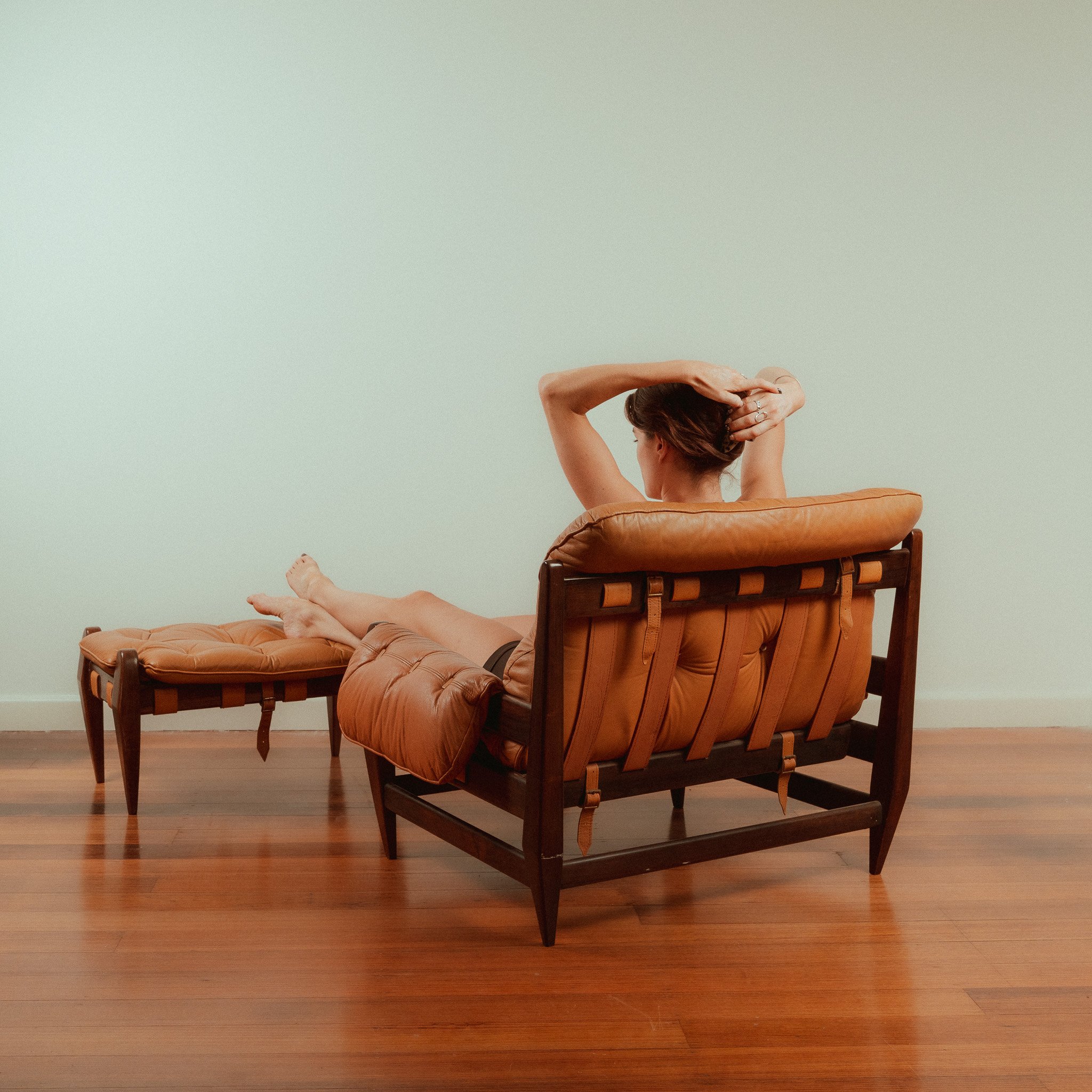A woman sitting on a leather chair with a matching ottoman, stretching her arms behind her head, in a room with hardwood floors and a plain white wall.