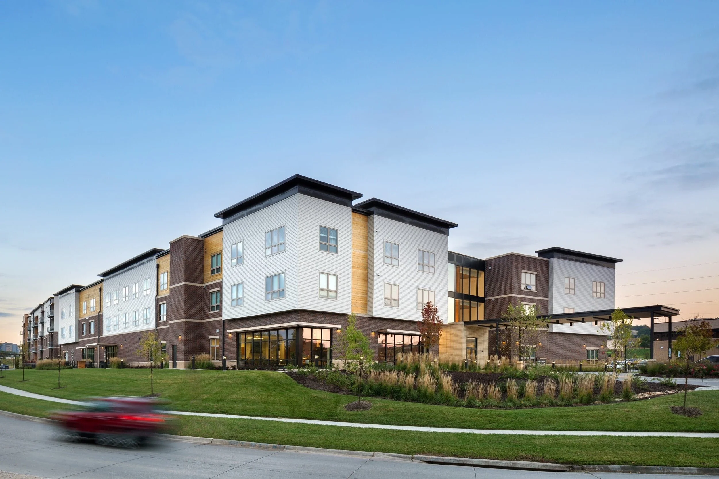 Modern multi-story apartment building with brick and white siding, large windows, landscaped lawn, and a blurred red car in motion on the road in front.