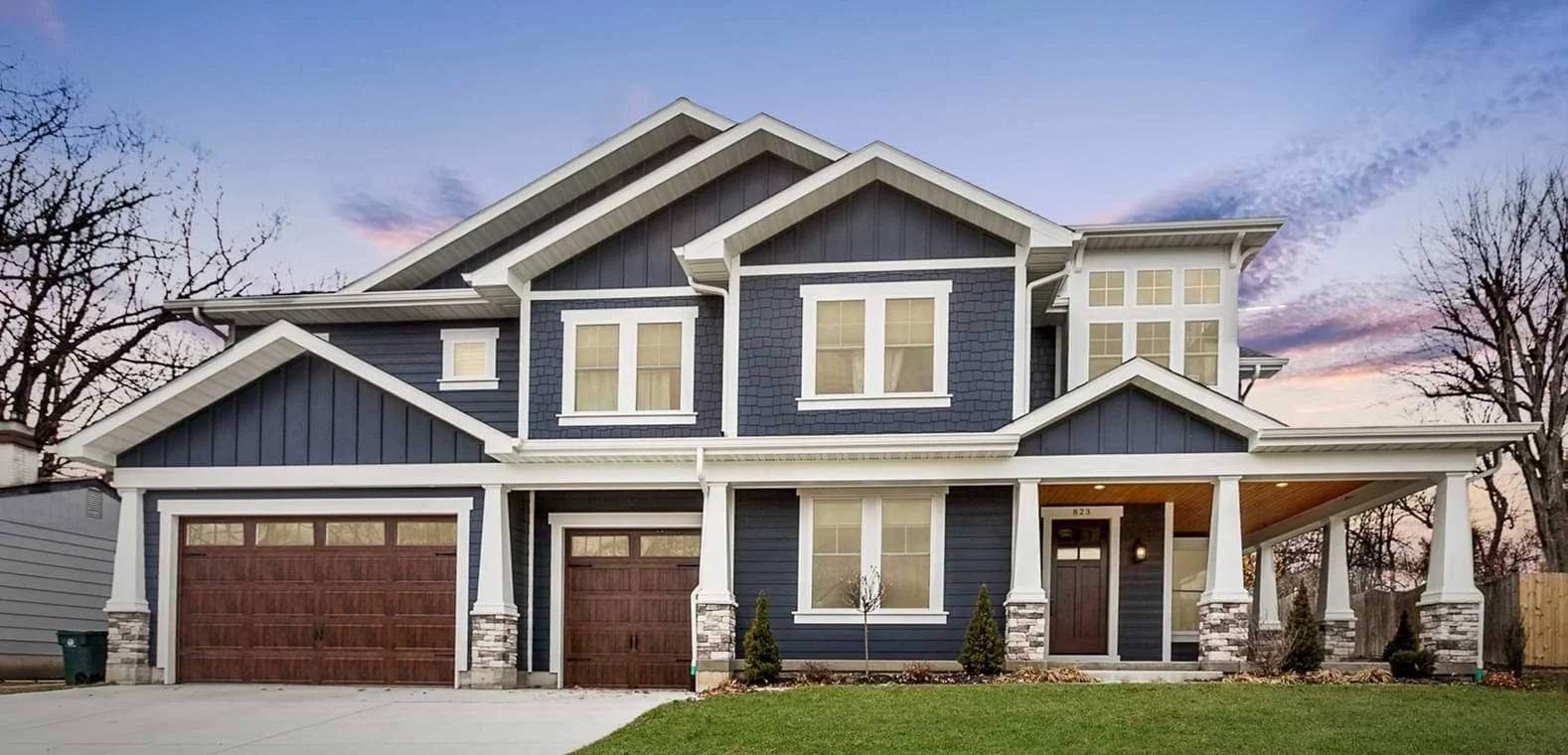 A two-story house with dark blue siding, white trim, and stone accents. It features a covered front porch, three garage doors, and multiple windows. The front yard has grass and small shrubs, with a clear sky in the background.