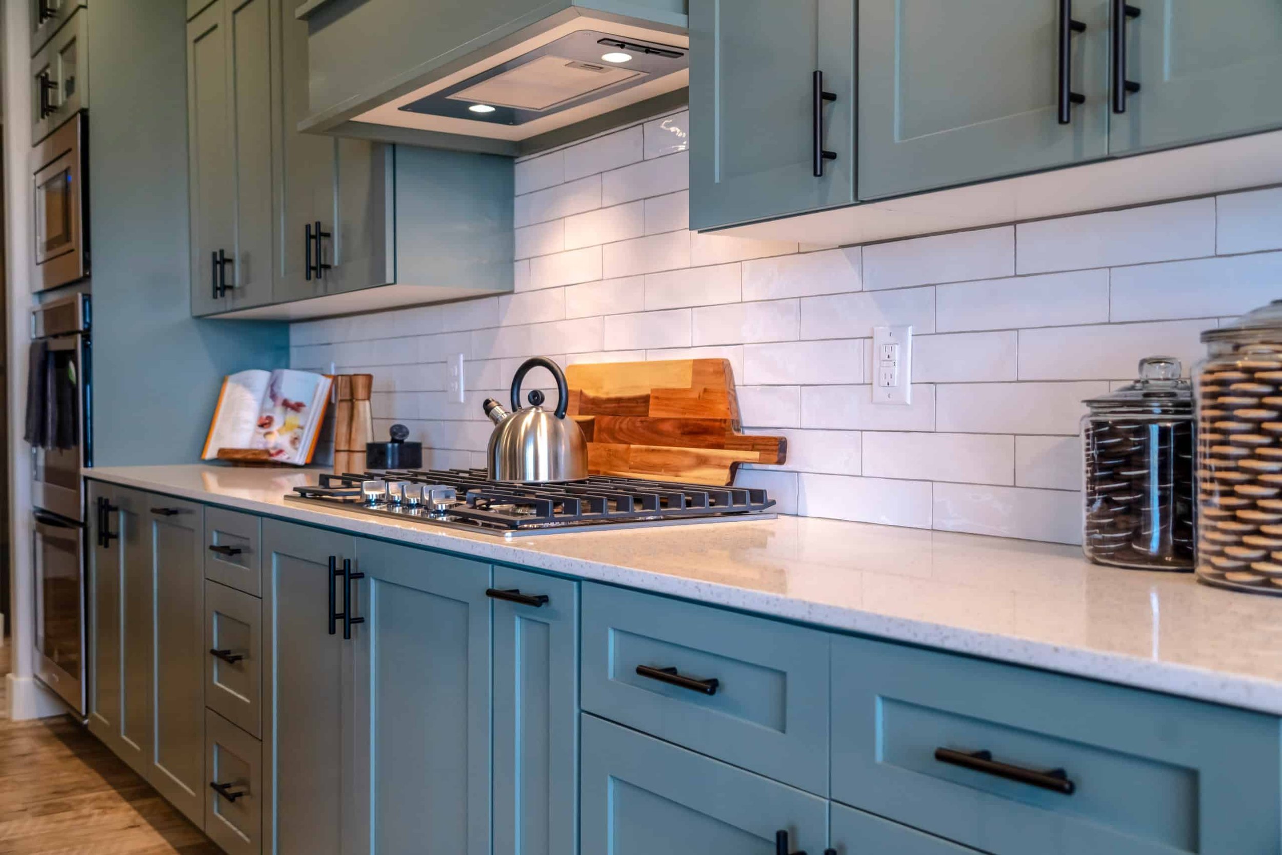 Modern kitchen with light blue cabinets, white countertop, white subway tile backsplash, stainless steel kettle on stove, wooden cutting boards, open cookbook, and glass jars with dry ingredients.