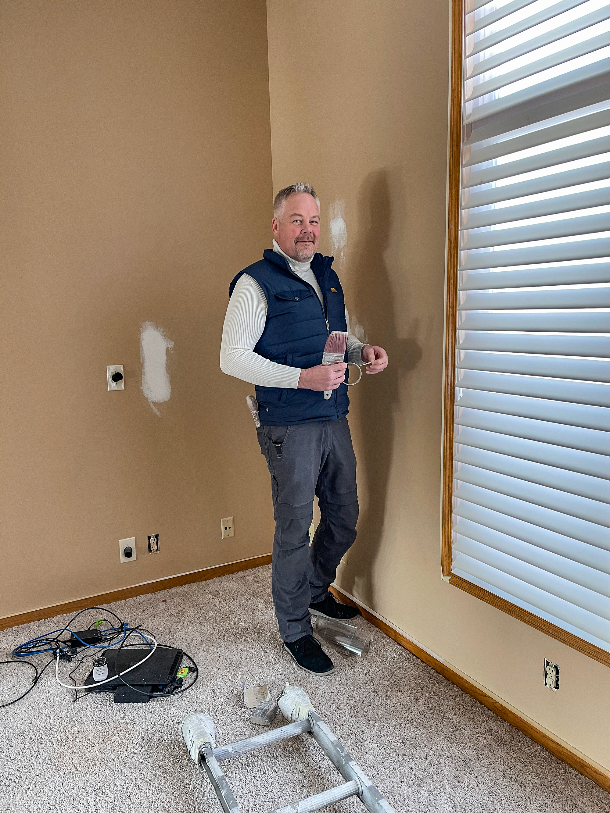 A man standing in a room under renovation, holding a paint roller, with patches of primer on the wall behind him, a ladder on the carpeted floor, and window blinds to his right.