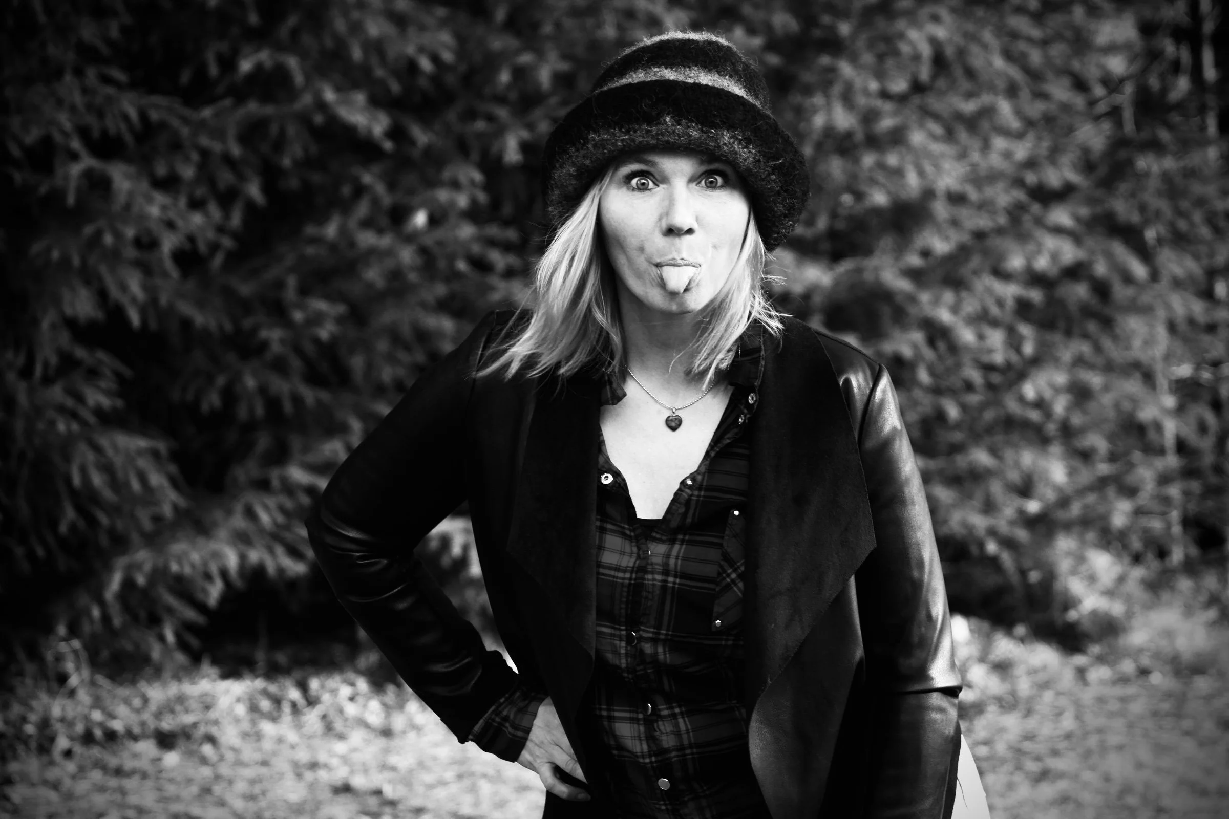 Junge Frau macht ein lustiges Gesicht, streckt die Zunge heraus und trägt einen Hut und eine Lederjacke, auf einem Waldweg im Freien.