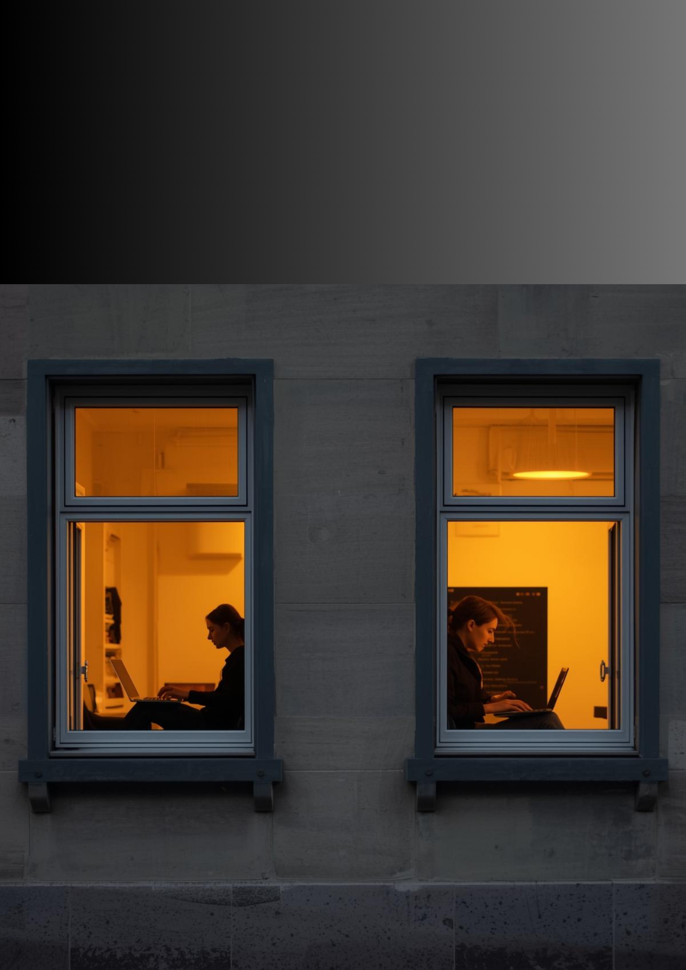 Zwei Frauen arbeiten abends in zwei gegenüberliegenden Fenstern eines Gebäudes, das mit grauem Beton verkleidet ist. Das Licht im Innenraum ist warm orange.