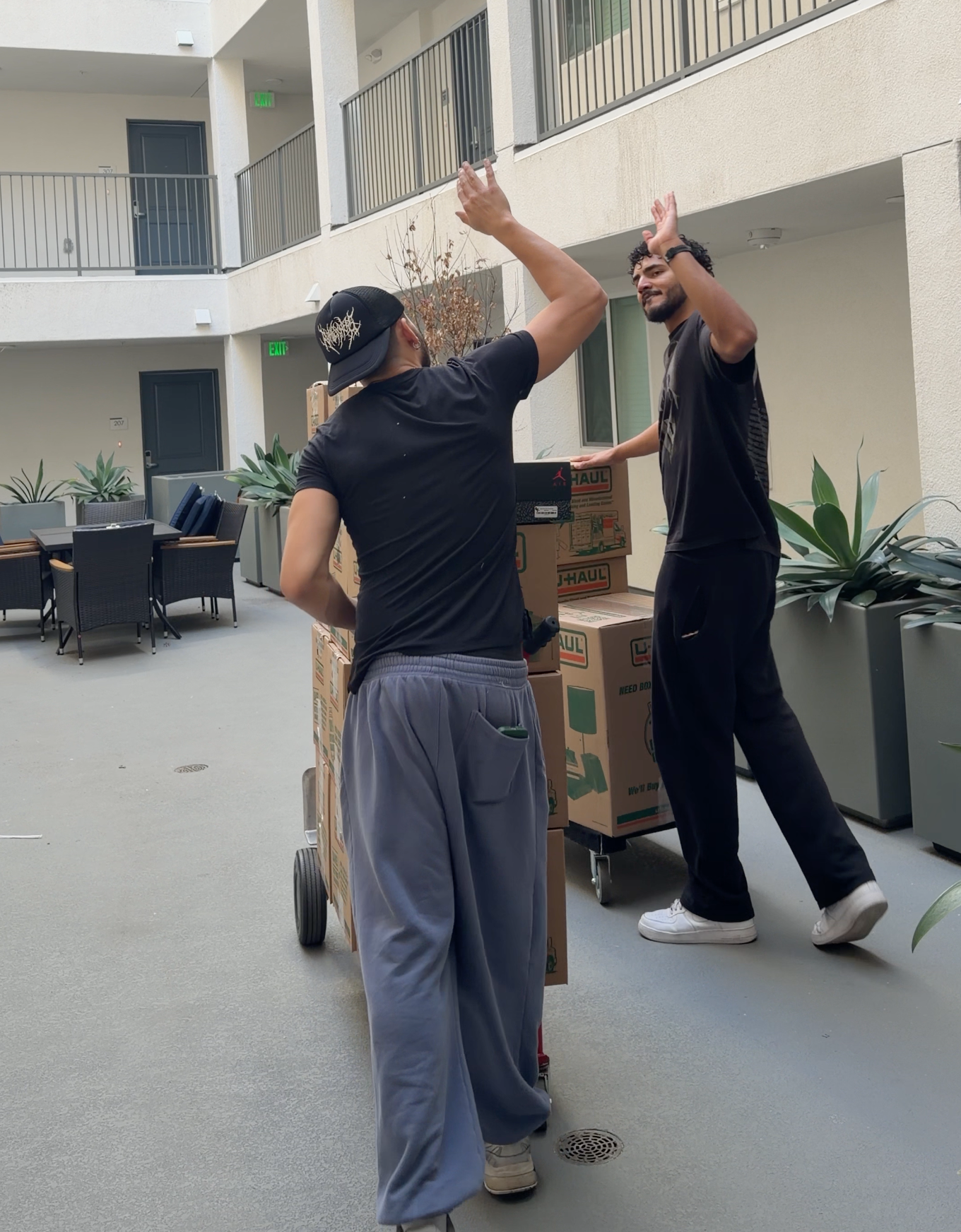 Two young men high-fiving in an apartment courtyard next to a cart with packed moving boxes.