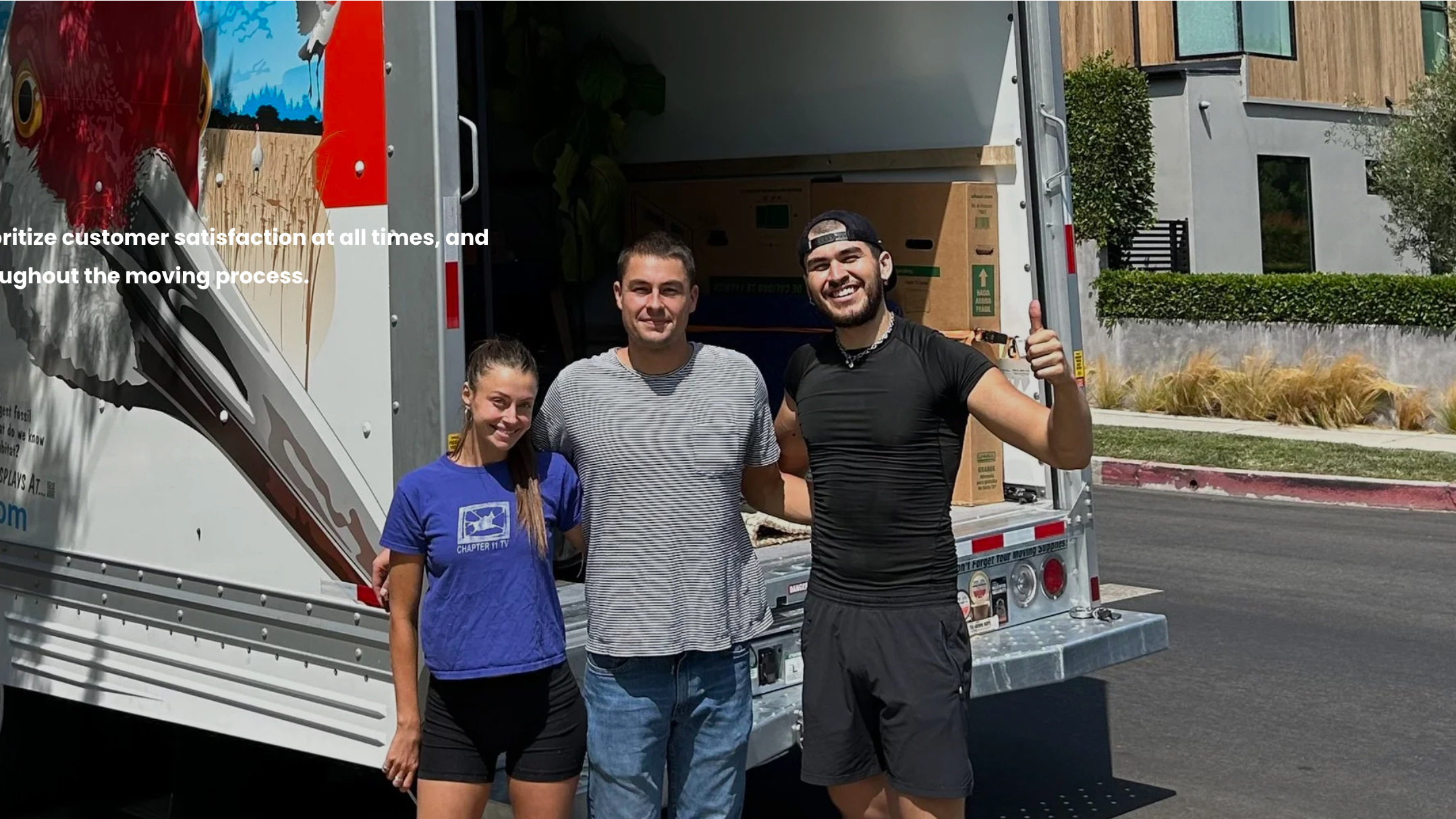 Three young adults standing outside a moving truck, smiling, with the truck open behind them, in a suburban neighborhood with modern houses and landscaping.