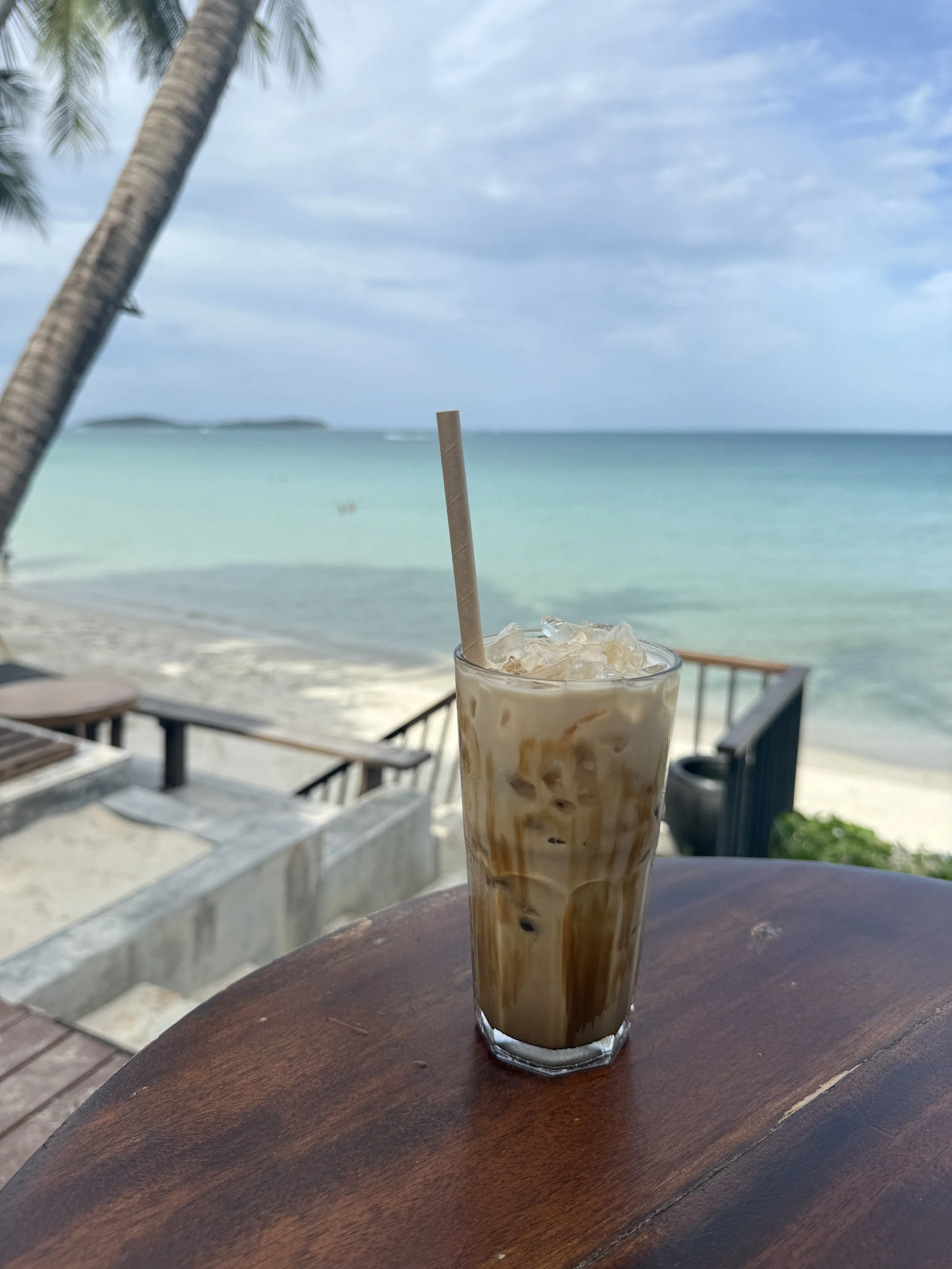 A thai iced coffee on a wooden table with a beach and ocean in the background.